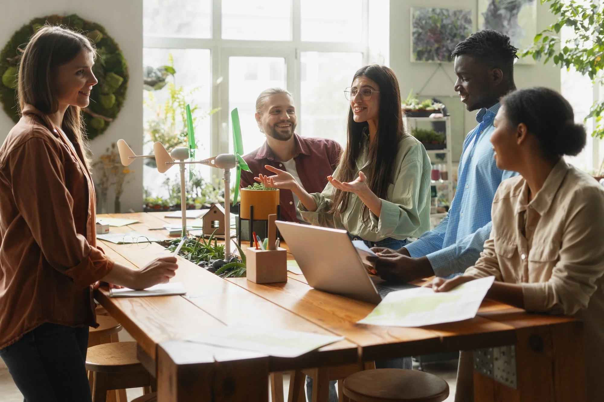 Groupe de cinq personnes discutant dans une salle lumineuse avec des plantes, une table en bois avec des ordinateurs, des plans et des objets de décoration, une femme explique à un groupe lors d'une réunion.
