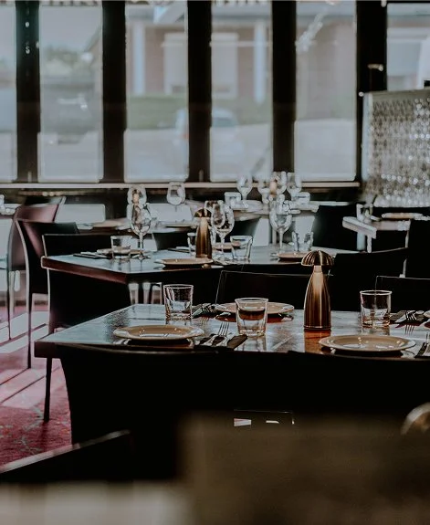 Elegant restaurant dining area with tables set for service, including plates, glasses, and silverware, near large windows.