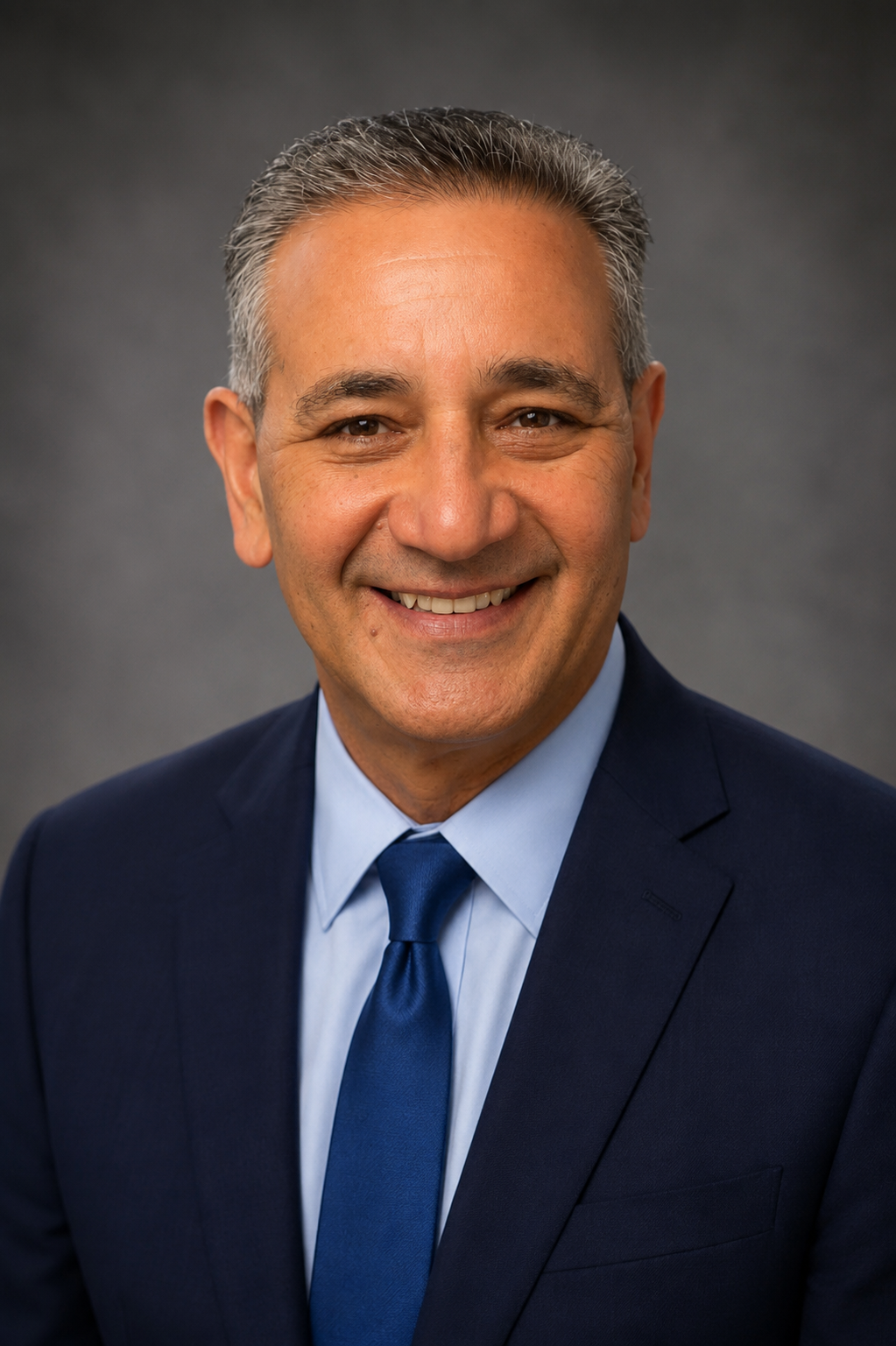 A professional headshot of Dr. Kelly Hanna wearing a dark navy suit, light blue shirt, and matching blue tie, smiling at the camera against a plain gray background.