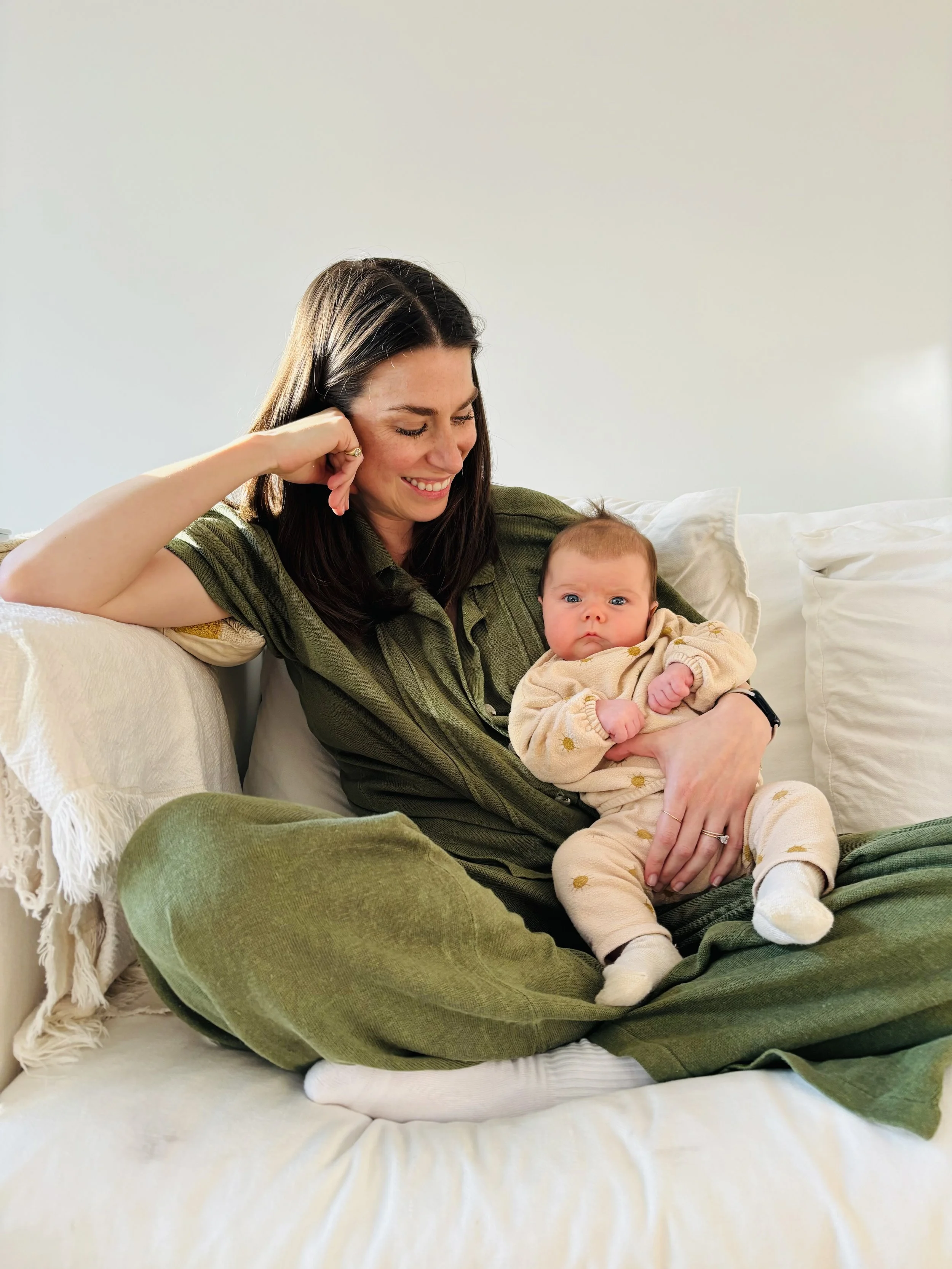 A woman with long dark hair sitting on a cream-colored couch, holding a baby with light brown hair and blue eyes in her lap. The woman is smiling and wearing a green shirt, while the baby looks curious. The background features a plain light wall and white pillows.