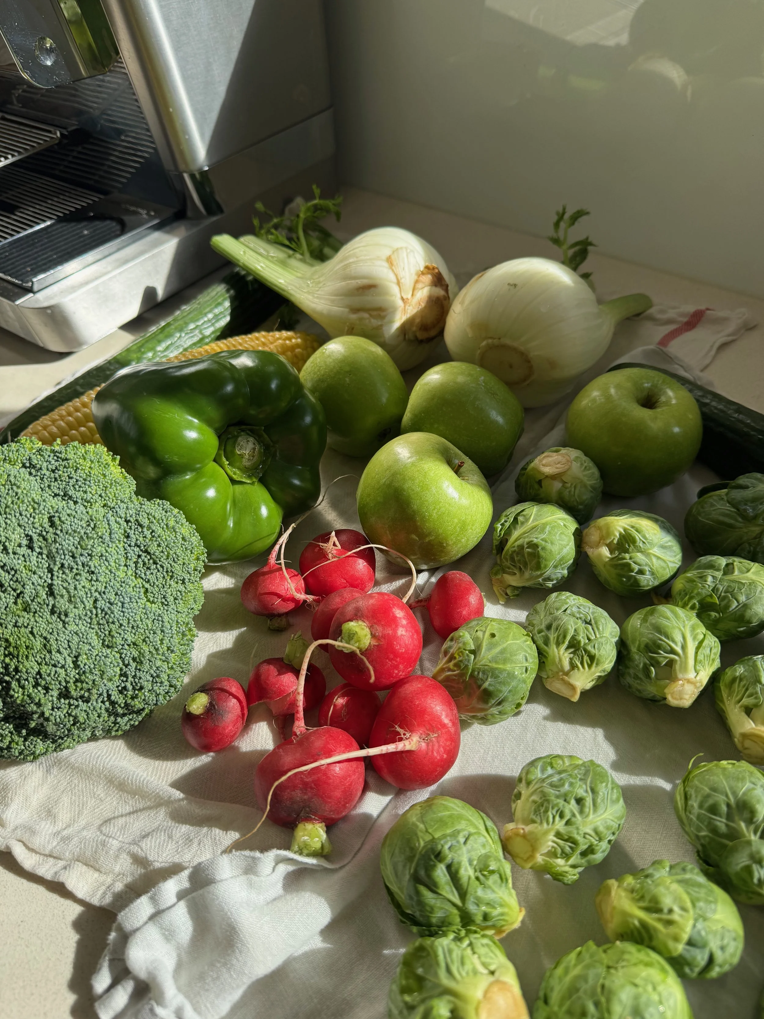 Fresh vegetables including broccoli, red radishes, green apples, brussels sprouts, cucumber, corn, and fennel on a white cloth and a countertop.