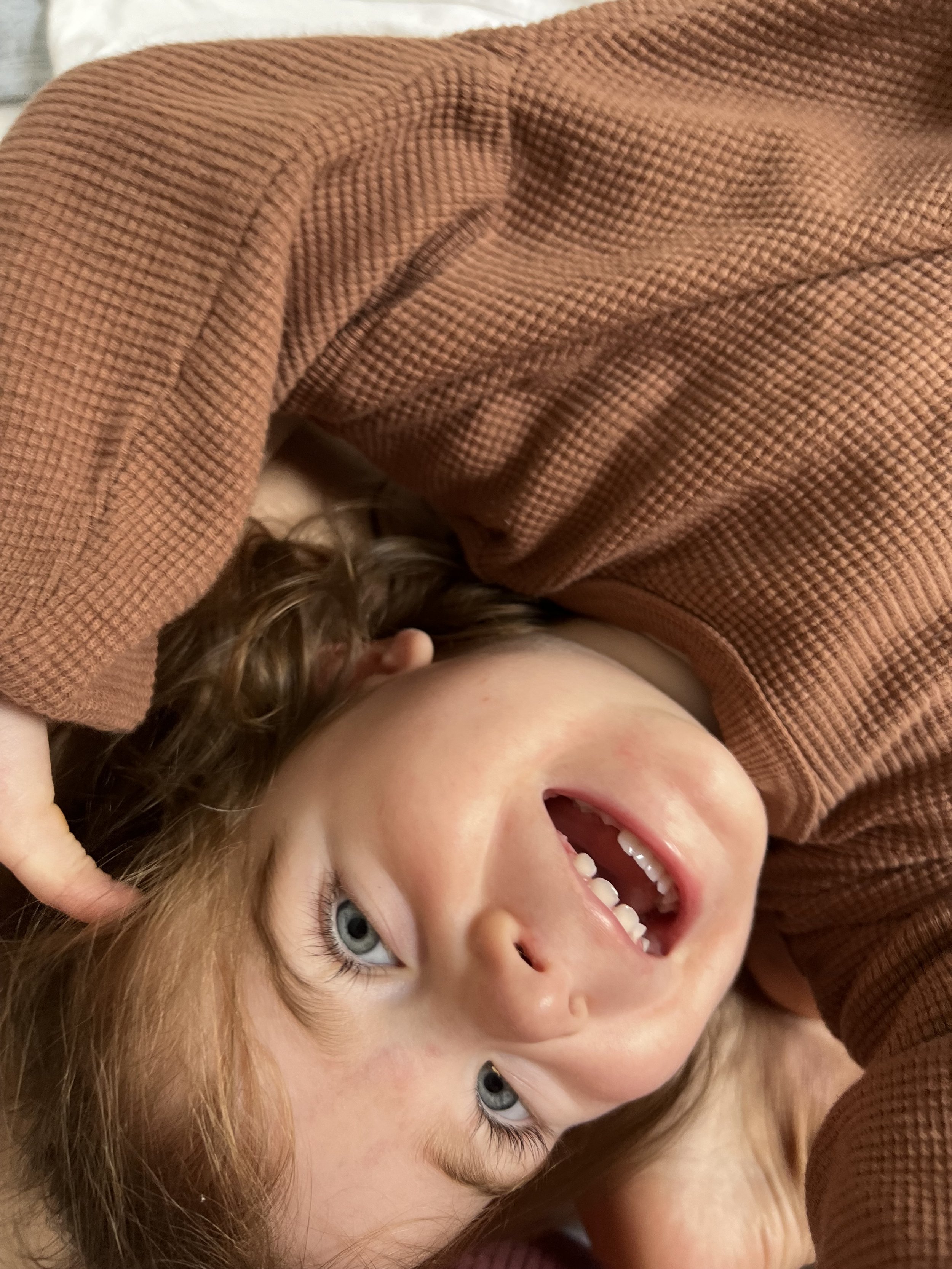 A young girl with blue eyes and red hair, smiling and lying down, wearing a brown top.