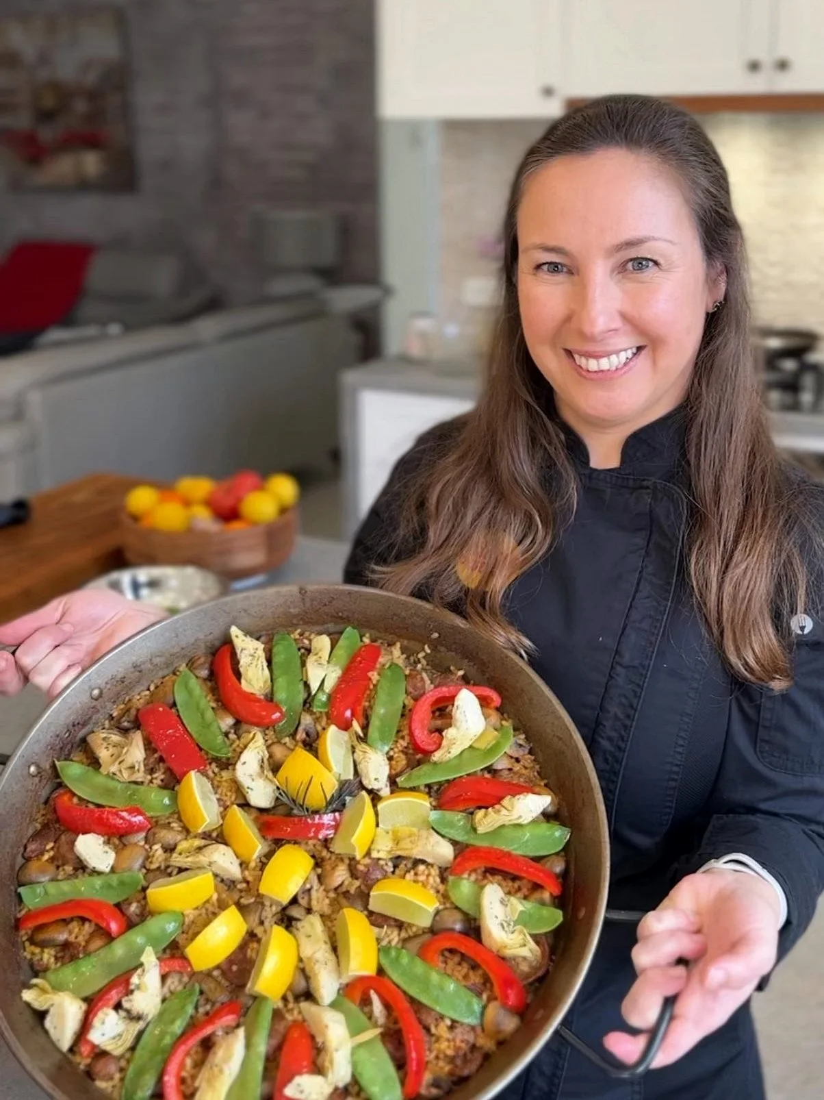 Portrait of Maria Vicencio Munchmeyer in a black shirt holding a large skillet of cooked food topped with red, yellow, and green bell peppers, and slices of cheese, with a smile on her face in a modern kitchen.