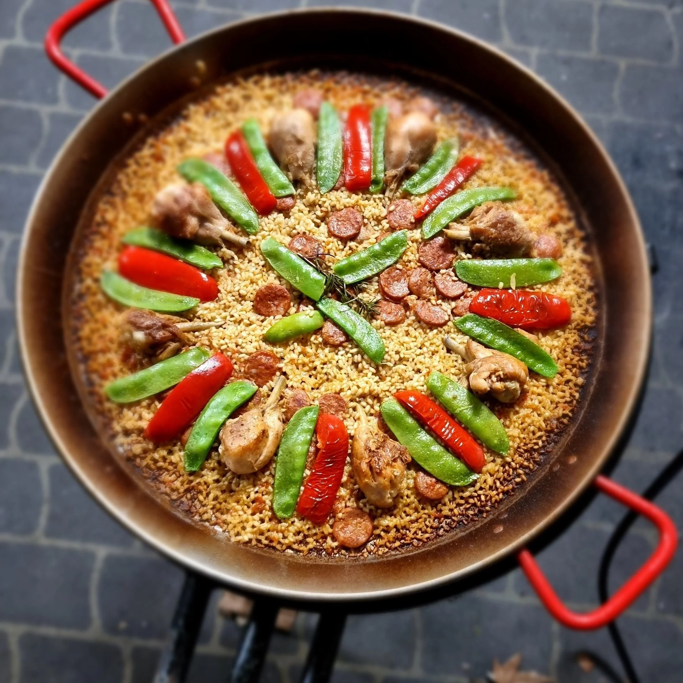 A black pan on a stovetop containing cooked grains with slices of red and green chili peppers, pieces of meat, and a sprig of herbs, with red stove burners visible in the background.
