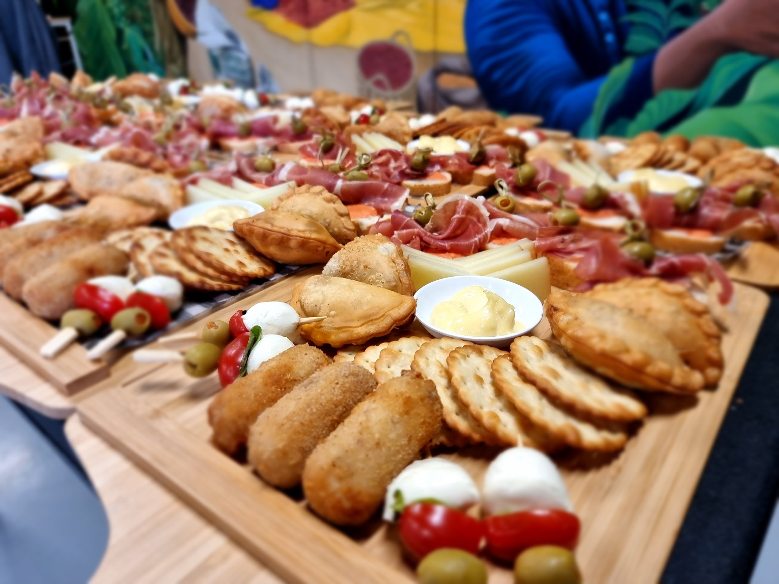 A wooden platter filled with various appetizers including slices of cured meats, cheese, small sandwiches, fried items, bowls of dip, and skewers with cherry tomatoes, mozzarella, and olives.
