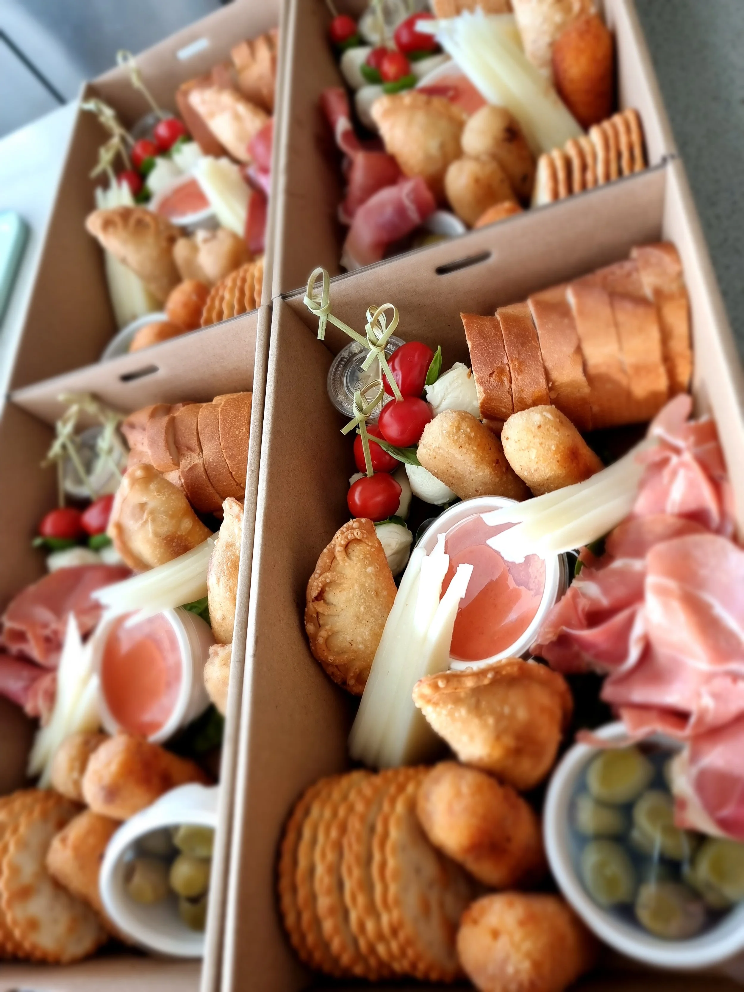 Three cardboard takeout boxes filled with assorted appetizers including cherry tomatoes, mozzarella cheese, prosciutto, fried cheese balls, crackers, and small salads.