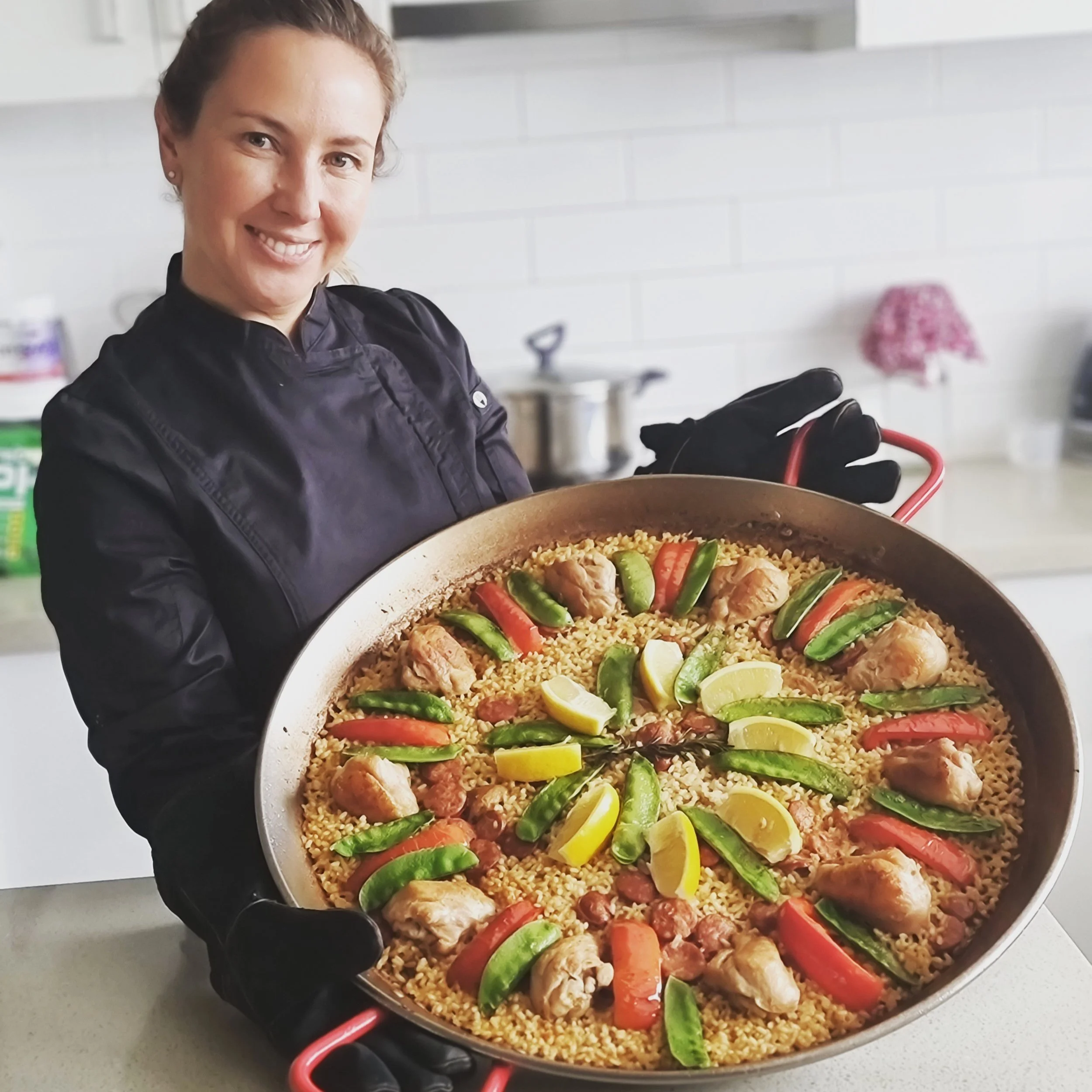Portrait of Maria Vicencio Munchmeyer, paella specialist, smiling while holding a large pan of cooked chicken and vegetable paella, in a kitchen setting.