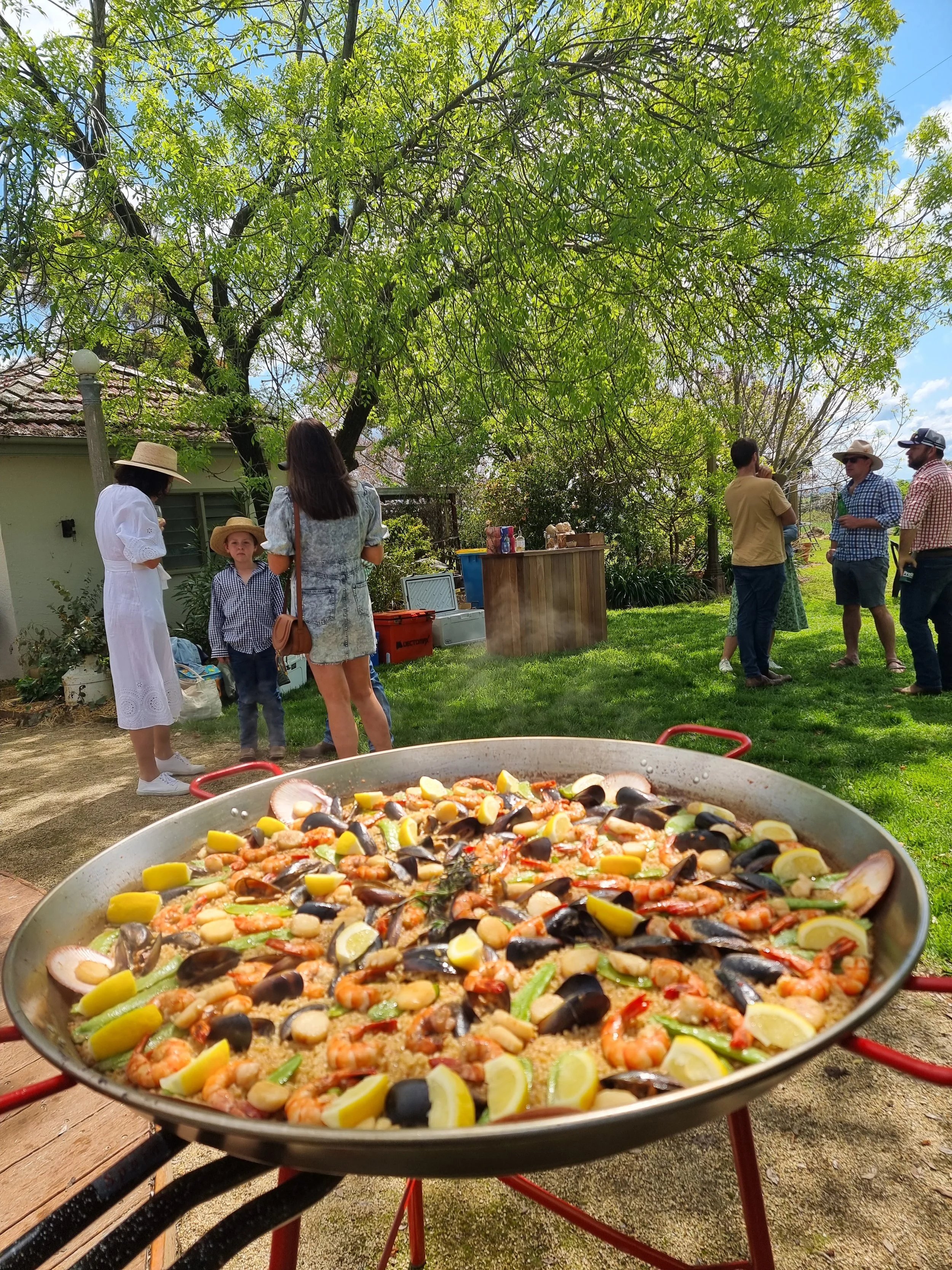 A large skillet filled with a seafood paella dish featuring shrimp, mussels, and vegetables, located outdoors on a sunny day.