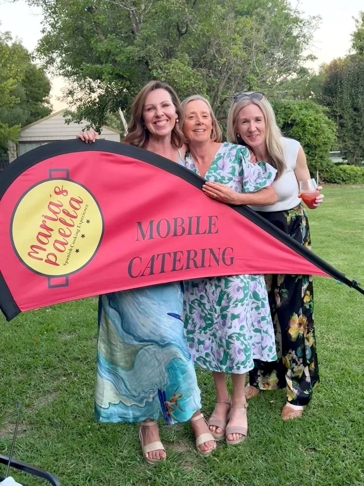 Three smiling women standing outdoors behind a red and black flag sail that reads "Maria's Paella Mobile Catering". One woman is holding a drink. They are dressed in summer clothes, standing on grass with trees and a house in the background.