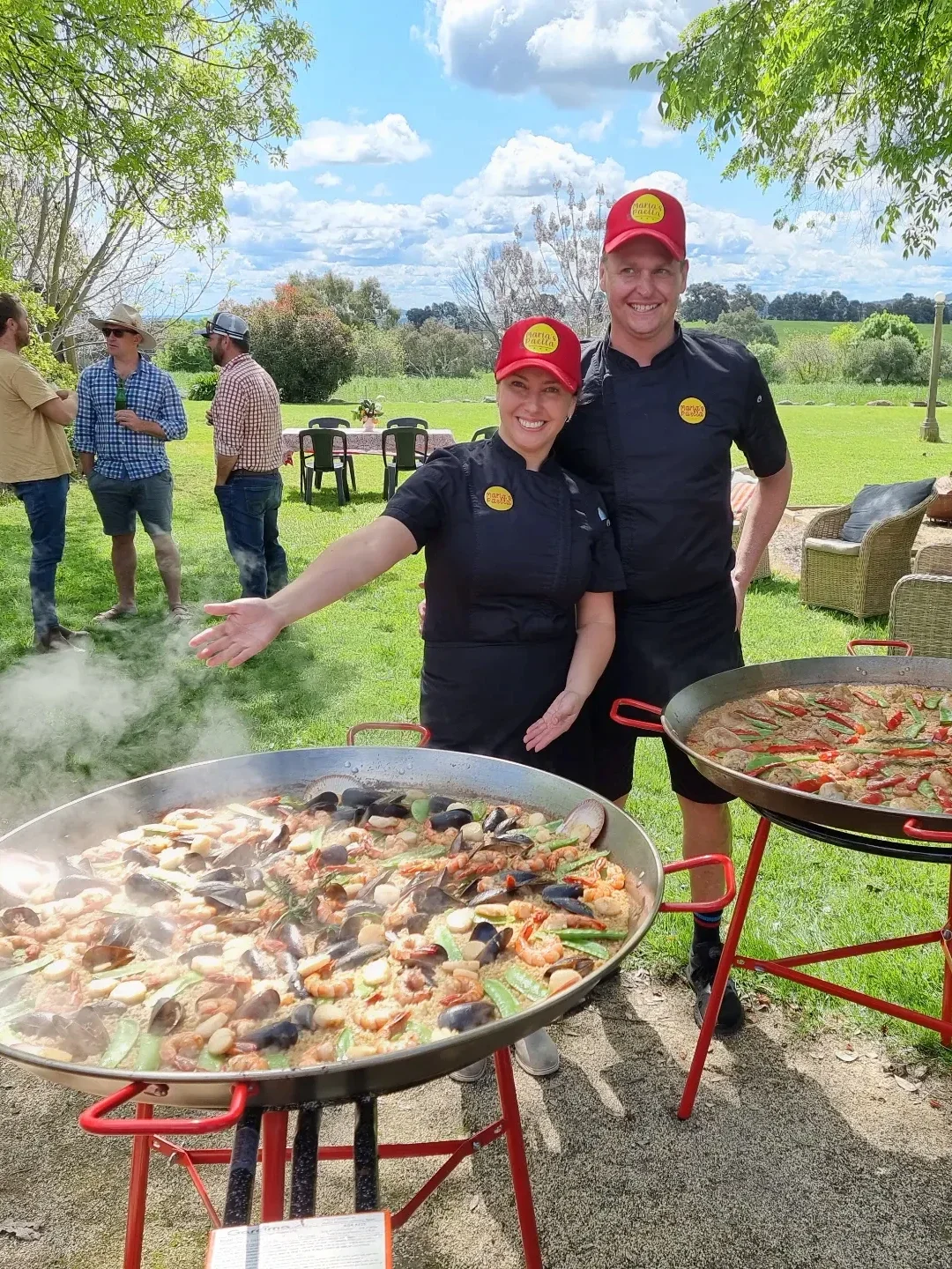 Two chefs smiling at an outdoor event with large pans of seafood paella cooking on red tripods, one woman gesturing towards it under a partly cloudy sky, with guests mingling in the background.