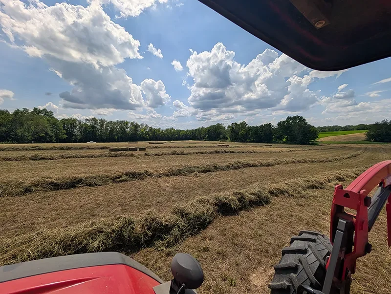 View from a tractor showing a field with cut hay and a blue sky with clouds, trees in the background.