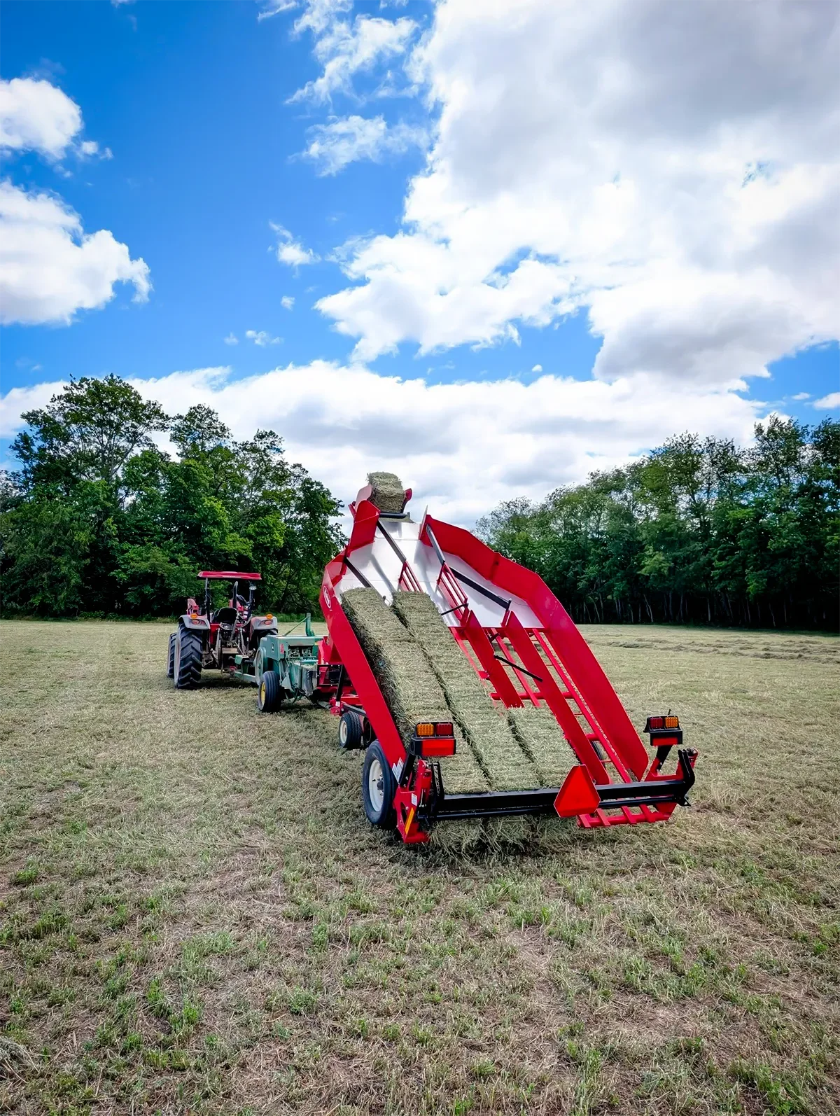 A red hay tedder attached to a green tractor working in a field, turning alfalfa under a partly cloudy sky with trees in the background.