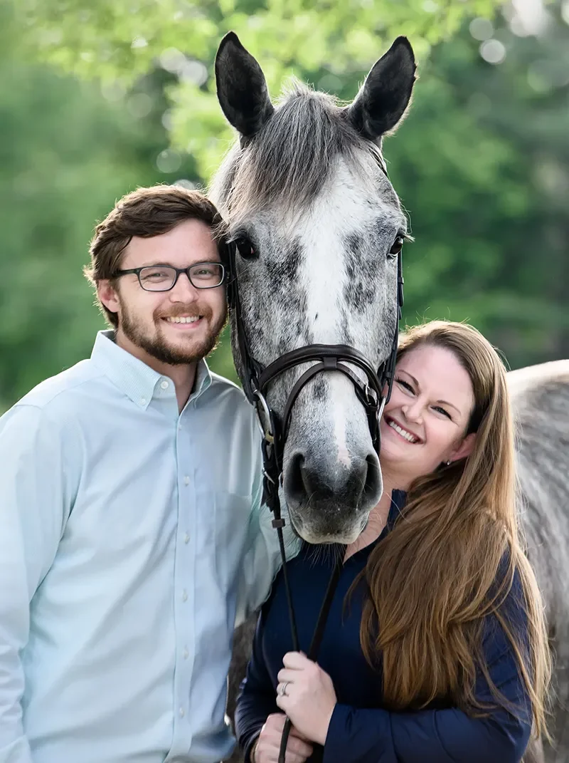 A smiling man and woman stand close together outdoors, holding a gray horse between them. The man is wearing glasses and a light blue shirt, the woman has long red hair and is dressed in a dark blue jacket.