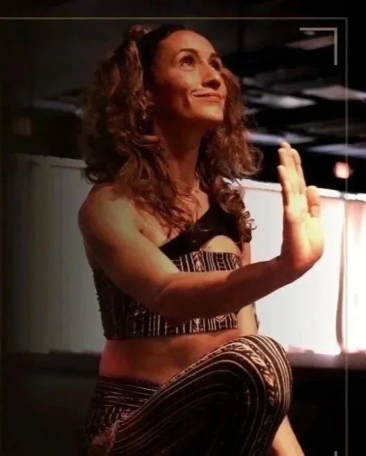 A woman with curly brown hair performing a yoga pose, kneeling with one hand raised and the other hand down, smiling softly in an indoor setting.