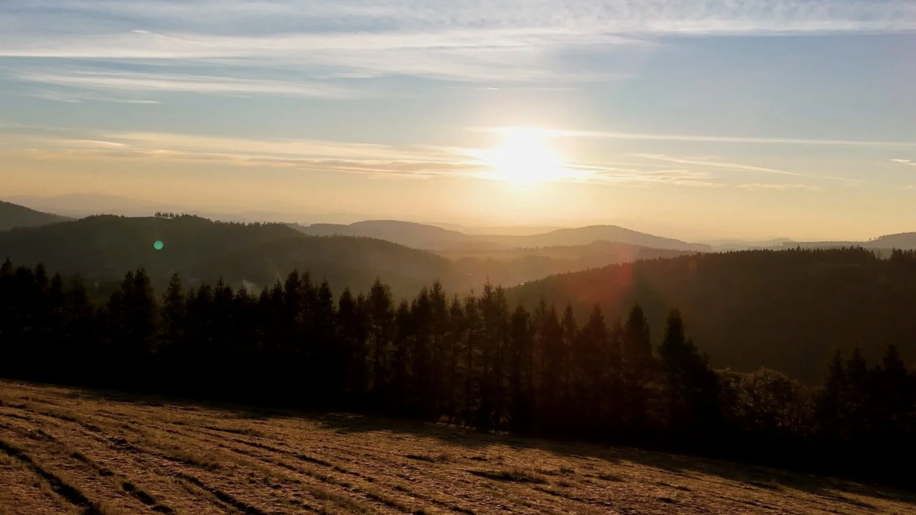 Sun setting over layered mountain ranges with a silhouette of trees in the foreground and a partly cloudy sky.