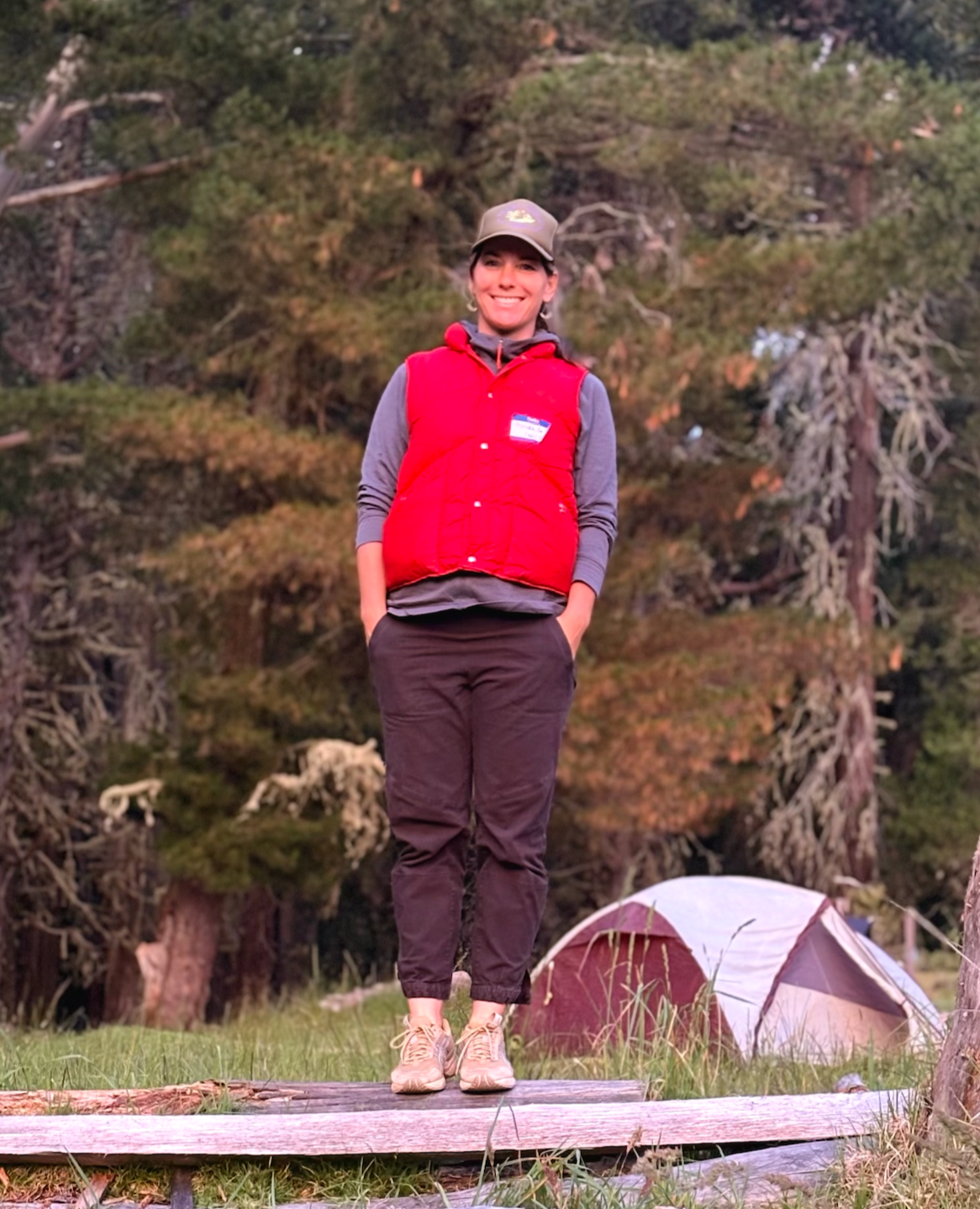 Maria standing outdoors in front of tall trees and a camping tent, wearing a red vest and a gray hoodie, smiling at the camera.