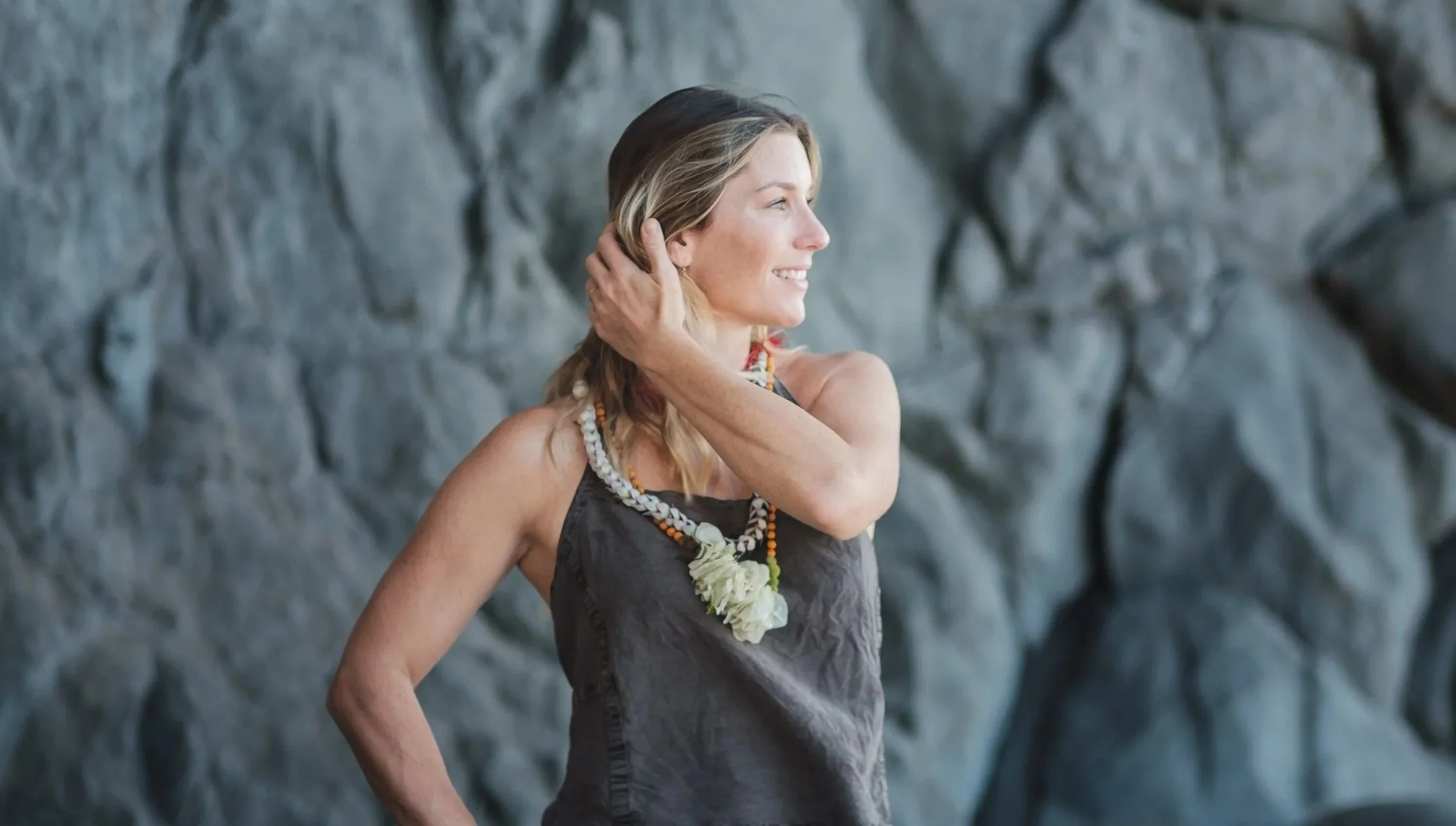 A woman with blonde hair wearing a dark gray dress and a shell necklace, smiling and touching her hair with her right hand, standing in front of a rocky background.