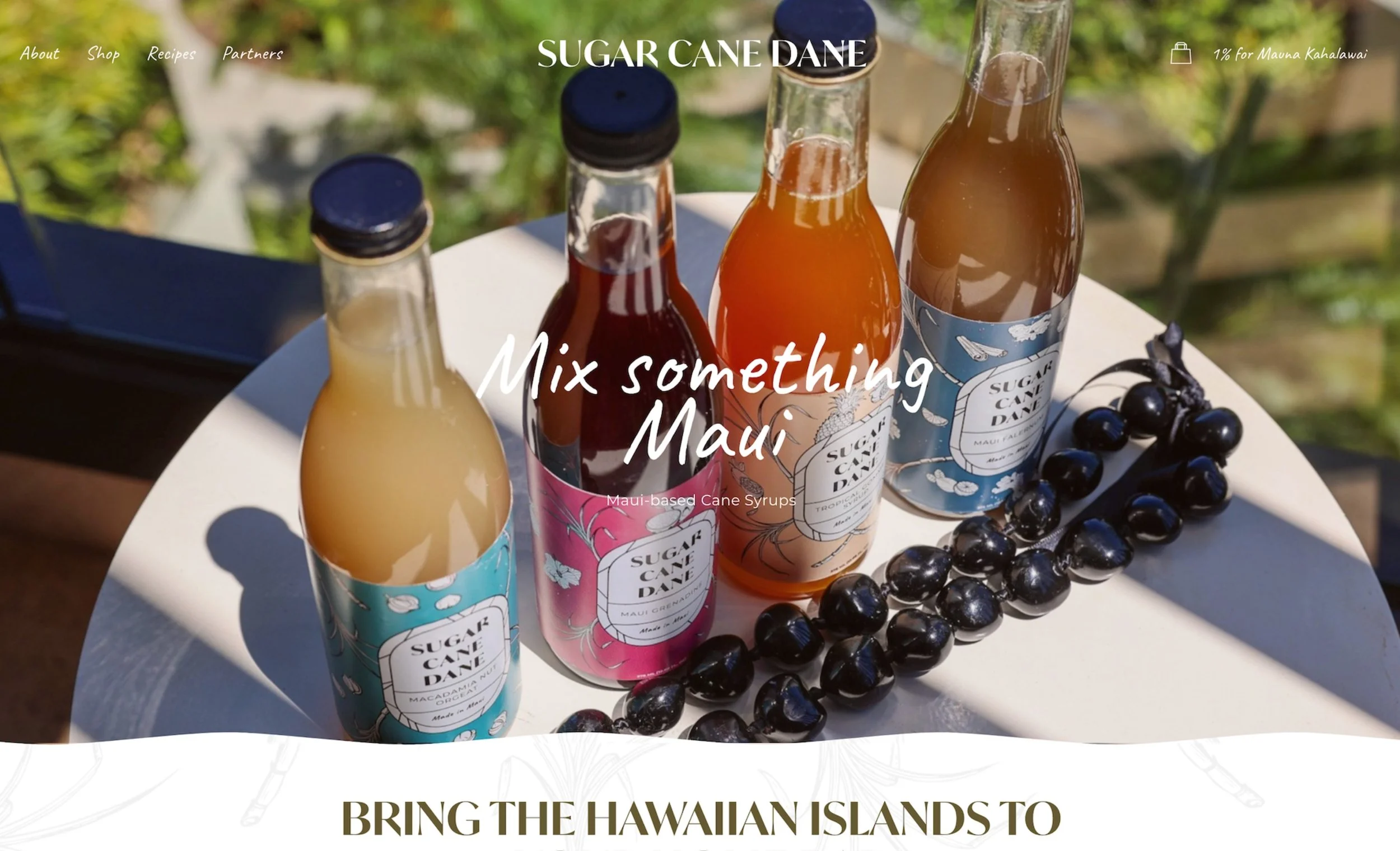 Assorted bottles of Hawaiian cane syrup with labels and a bunch of black cherries on the side, placed on a round white table outside with greenery in the background.