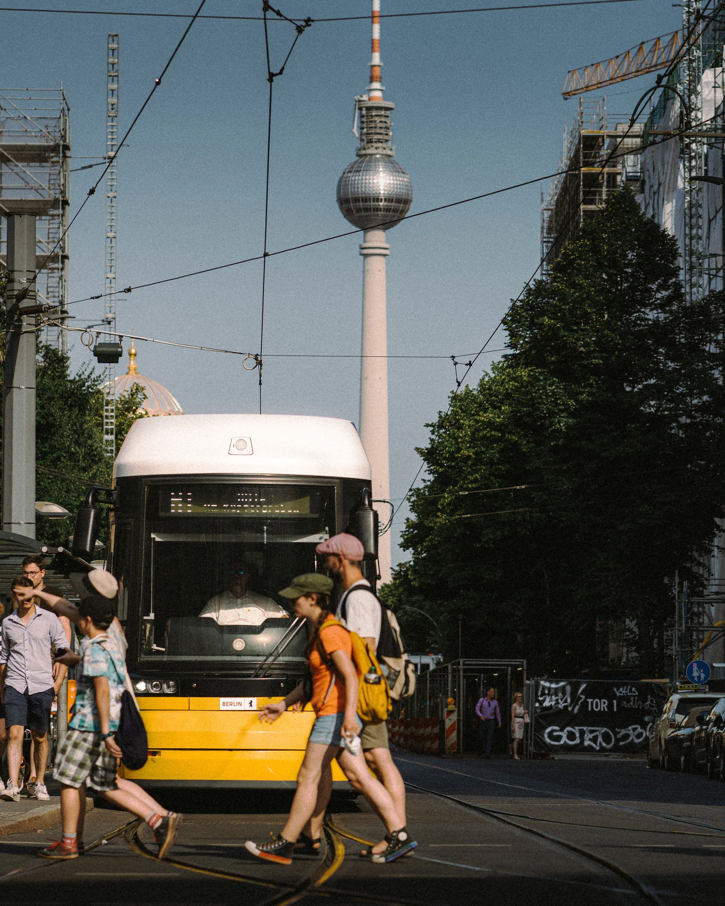 A yellow bus labeled Berlin, with people walking in front of it on a city street. In the background, the Berlin TV Tower is visible against a clear blue sky, surrounded by scaffolding and construction elements, with some graffiti on barriers.