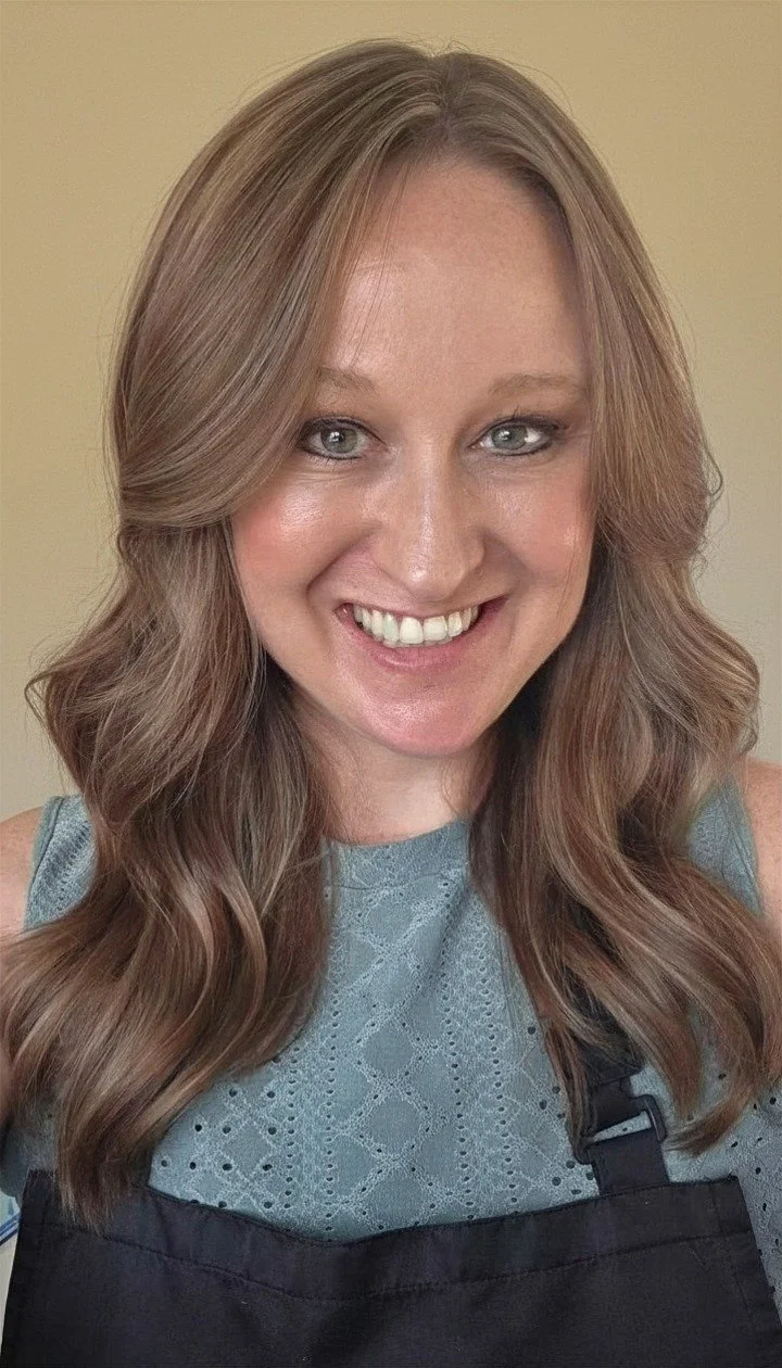 A woman with long, wavy, light brown hair and blue eyes smiling at the camera, wearing a gray top and a black apron, standing against a plain beige background.