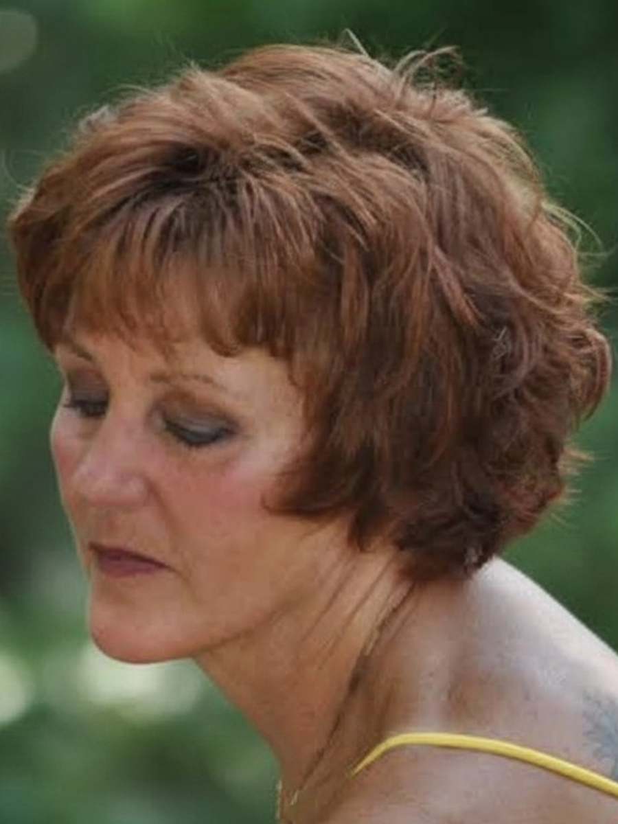 Side profile of a woman with short, wavy auburn hair and light skin, wearing a yellow strap top, with a blurred green background.