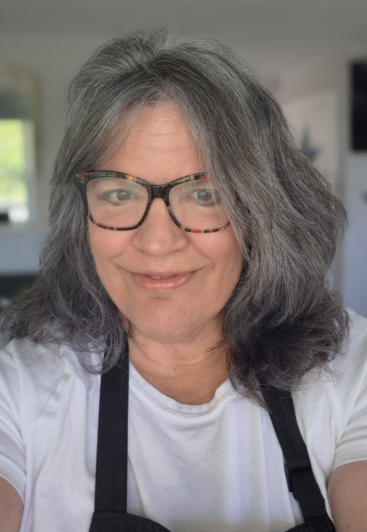 A woman with gray hair, glasses, and a white shirt smiling, with a blurred indoor background including a window and a television.