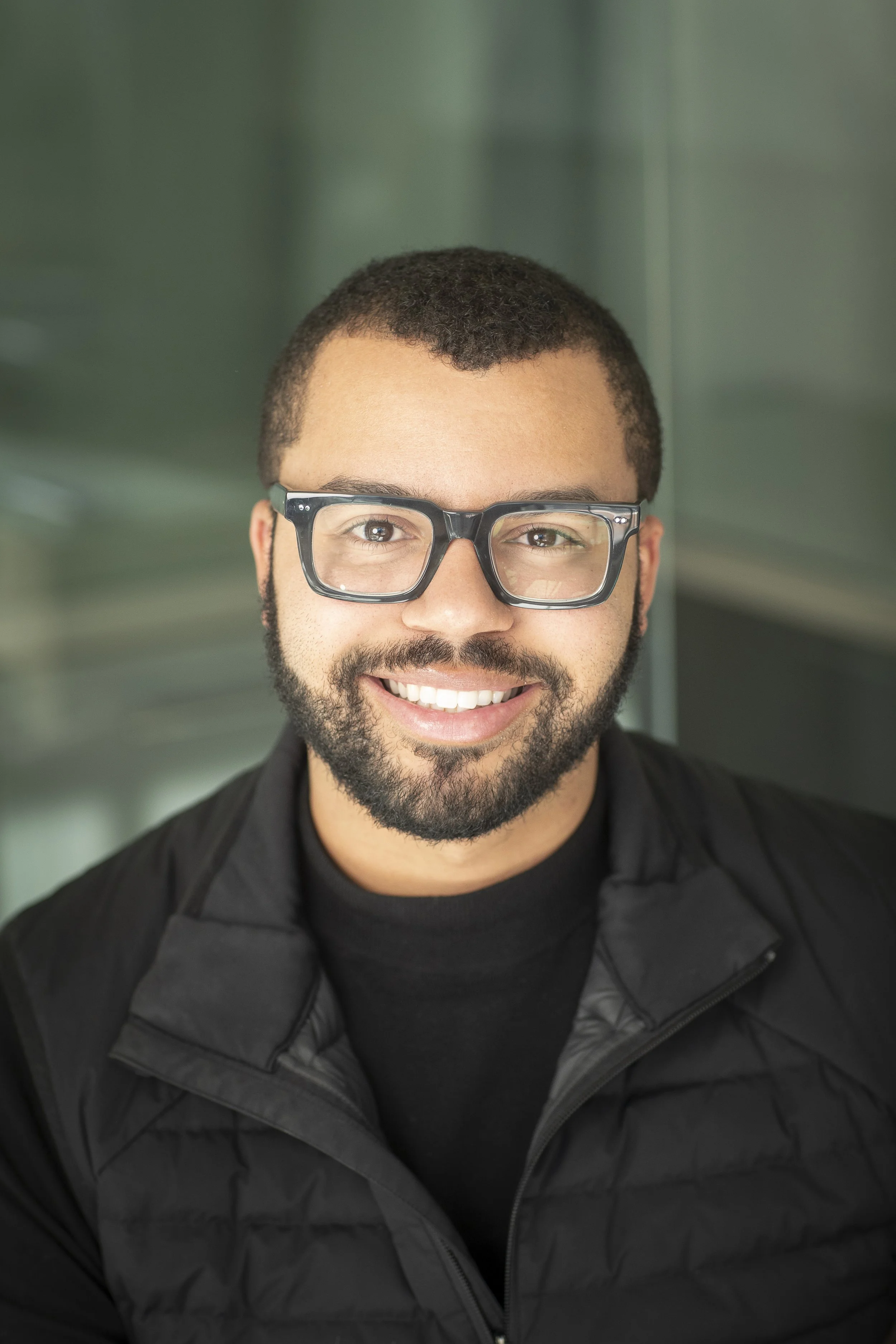 Close-up of a smiling man with short curly hair, a beard, glasses, wearing a black jacket, in front of a blurred background.