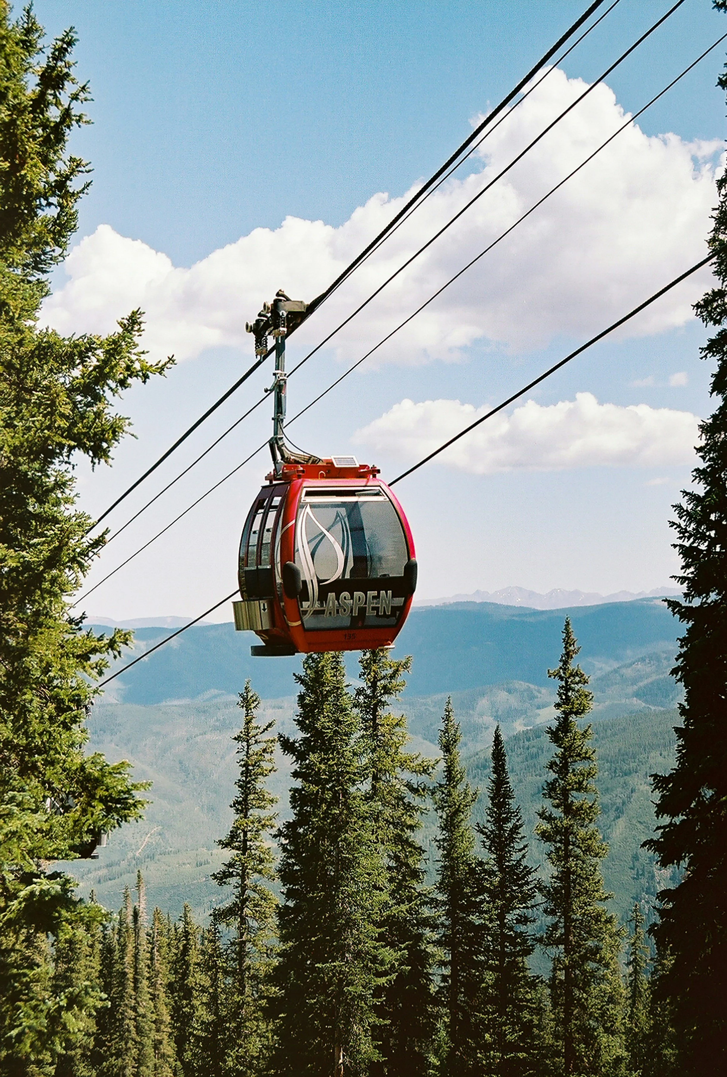 A red gondola lift traveling above a forested mountain landscape under a partly cloudy sky.