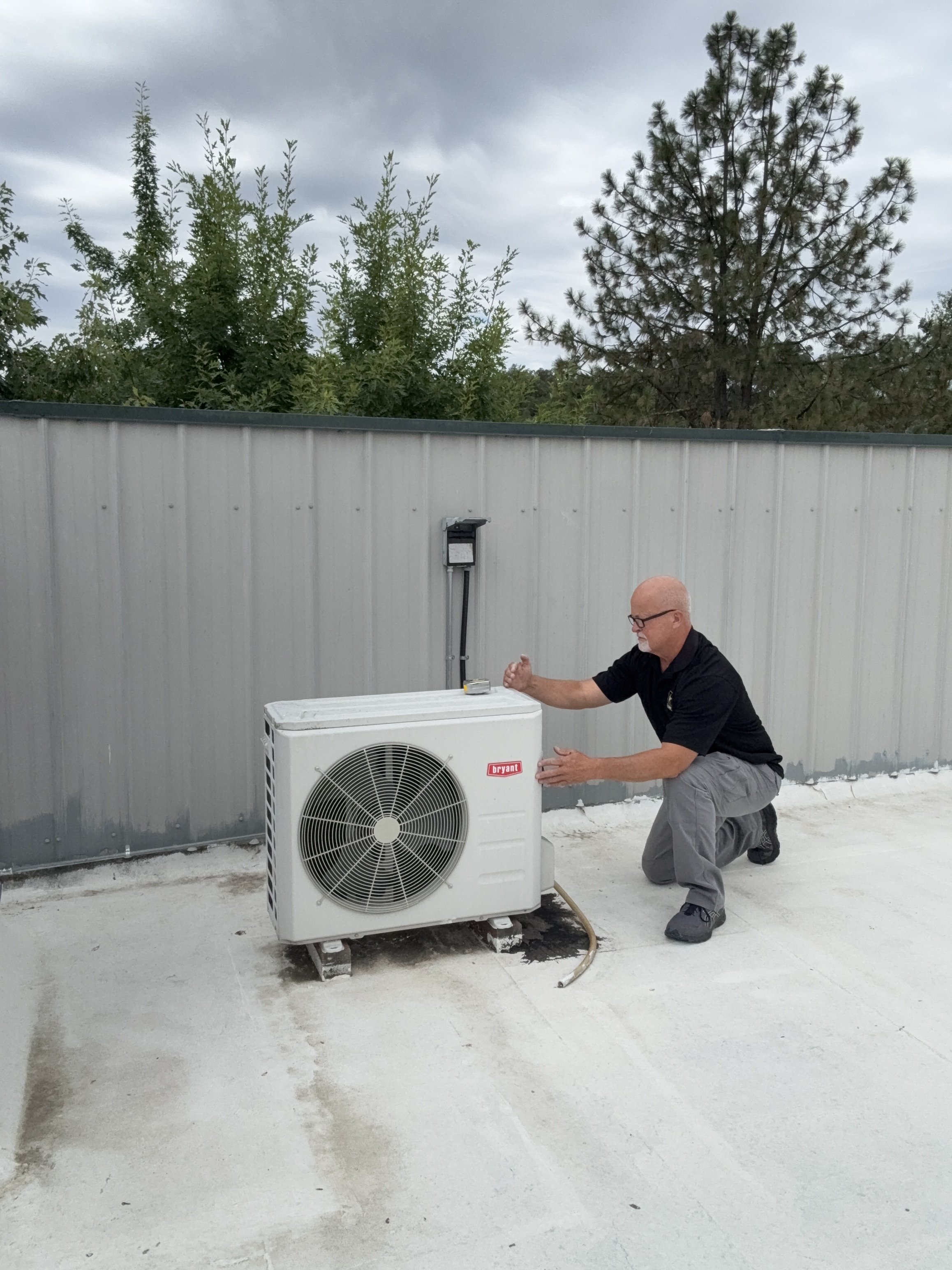 Man kneeling on the ground inspecting or installing an outdoor air conditioning unit on a flat surface with a metal wall and trees in the background.