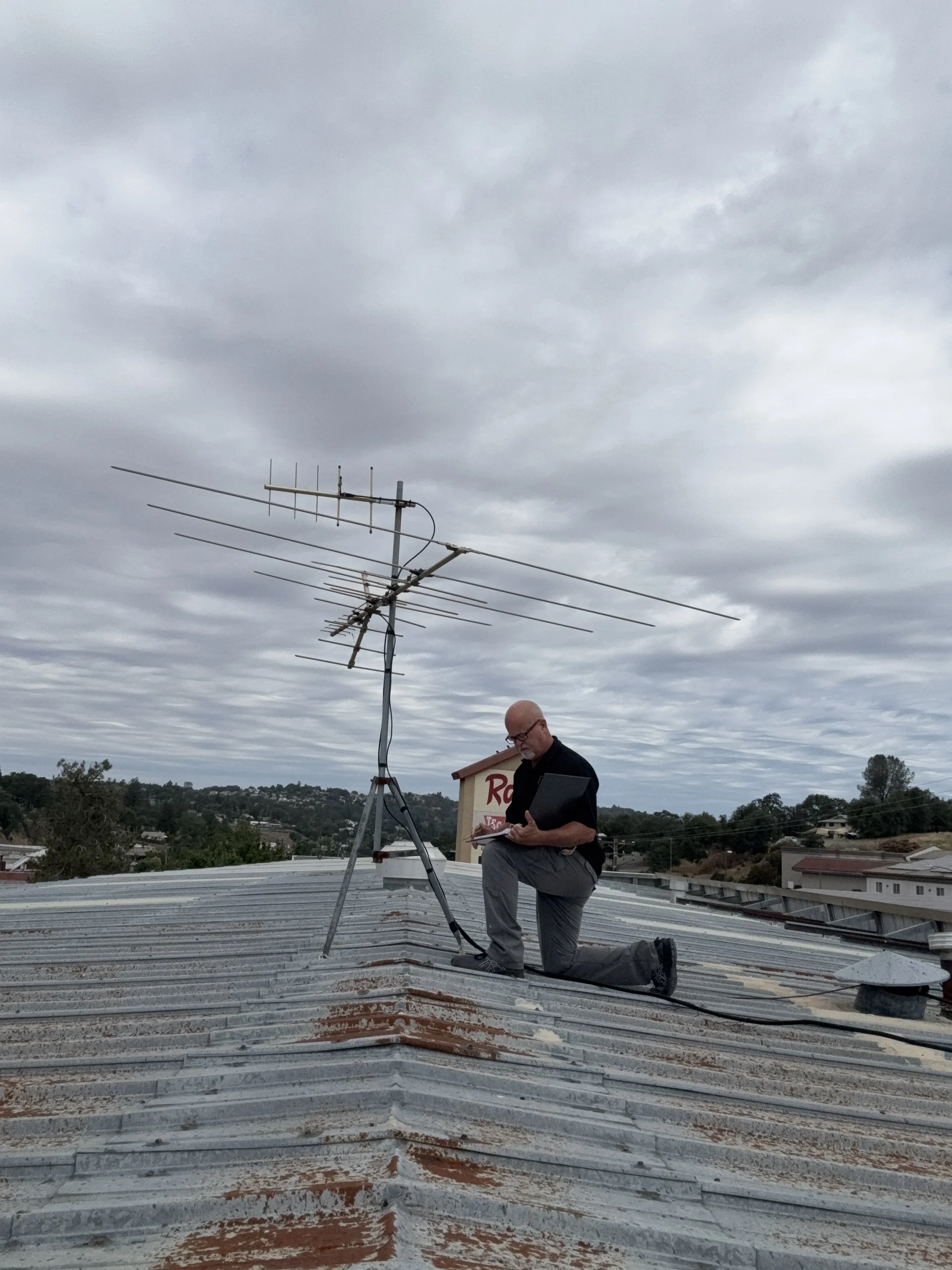 Man kneeling on a roof inspecting or working on antennas and electrical equipment under cloudy sky.