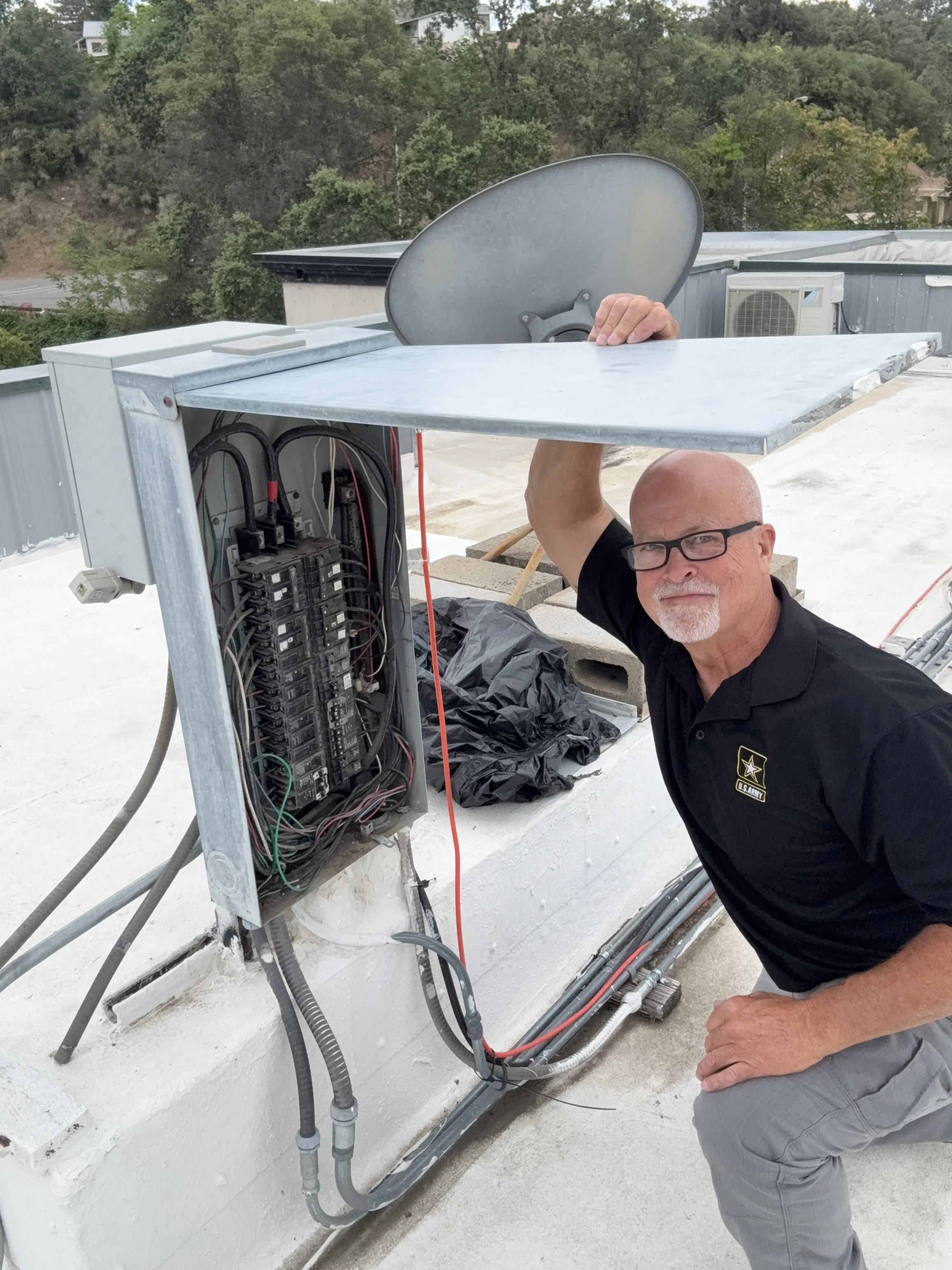 Man wearing glasses inspecting an outdoor electrical panel with wires and components, on a rooftop with trees and buildings in the background.