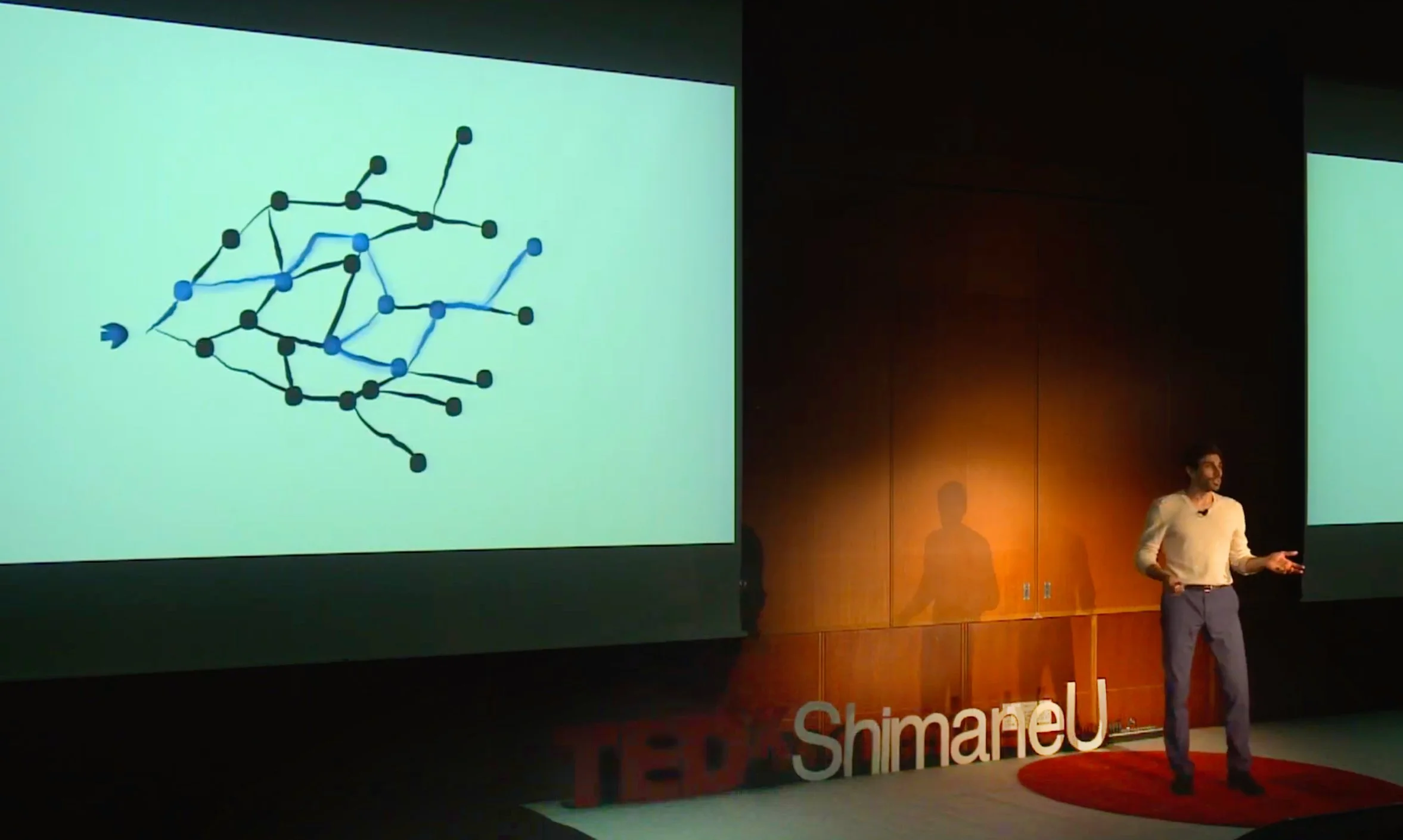 A male speaker presenting at a TEDxShimaneU event, with a large screen behind him displaying a diagram of interconnected nodes.