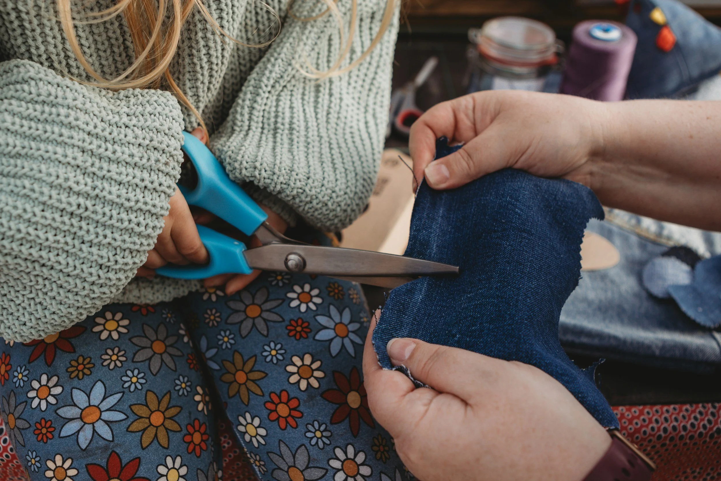 Someone is cutting someone's jeans with scissors while another person holds their leg. The person holding their leg is wearing a floral skirt. The person cutting is wearing a gray sweater. There are craft supplies in the background.