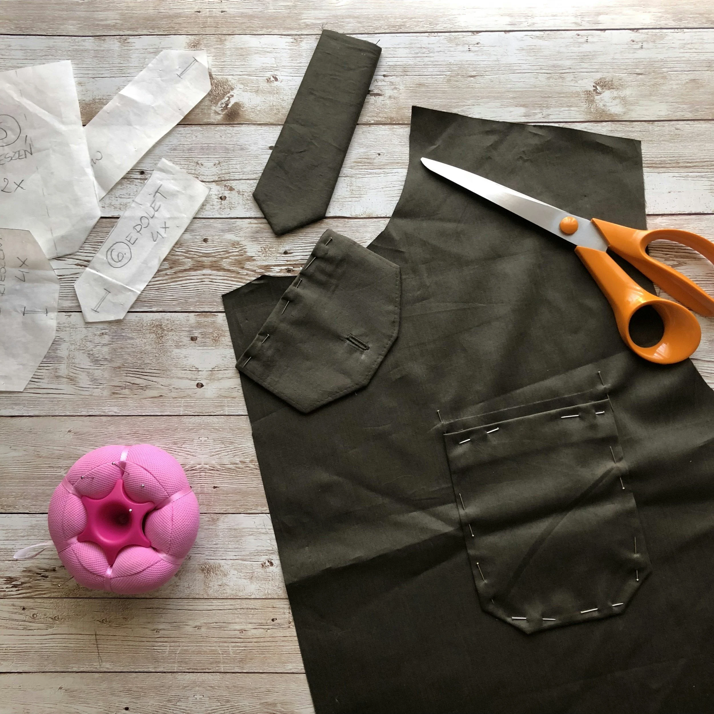 A pair of orange-handled scissors, black fabric, pink craft foam, white paper with handwritten notes, and a pink floral-shaped object on a wooden surface.