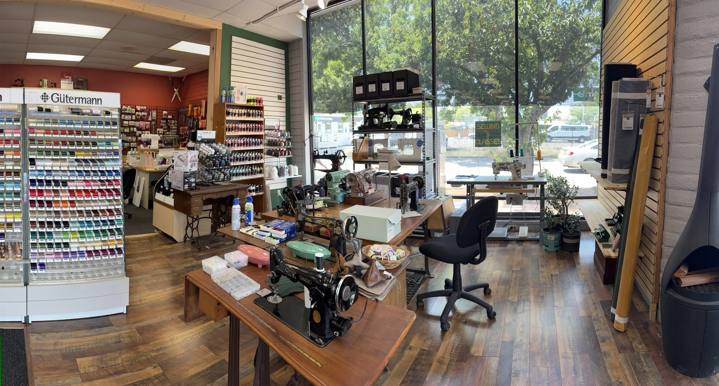 Interior of a craft or hobby store with sewing machines, shelves of thread, and craft supplies on wooden tables near large windows showing outside trees and parking lot.