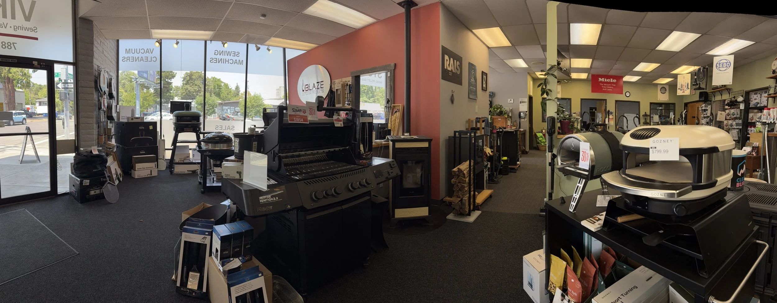 Indoor store selling appliances and home equipment, with a large front window showing outside parking lot and trees, and various appliances and products displayed on shelves and tables.