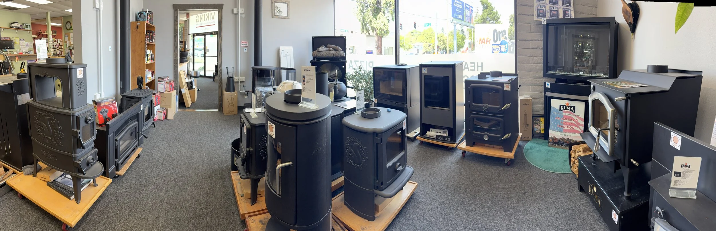 Store interior with wood stoves on display, including brands like Kuma, arranged on wooden platforms. There are large windows letting in natural light, with some home furnishings and electronics visible in the background.