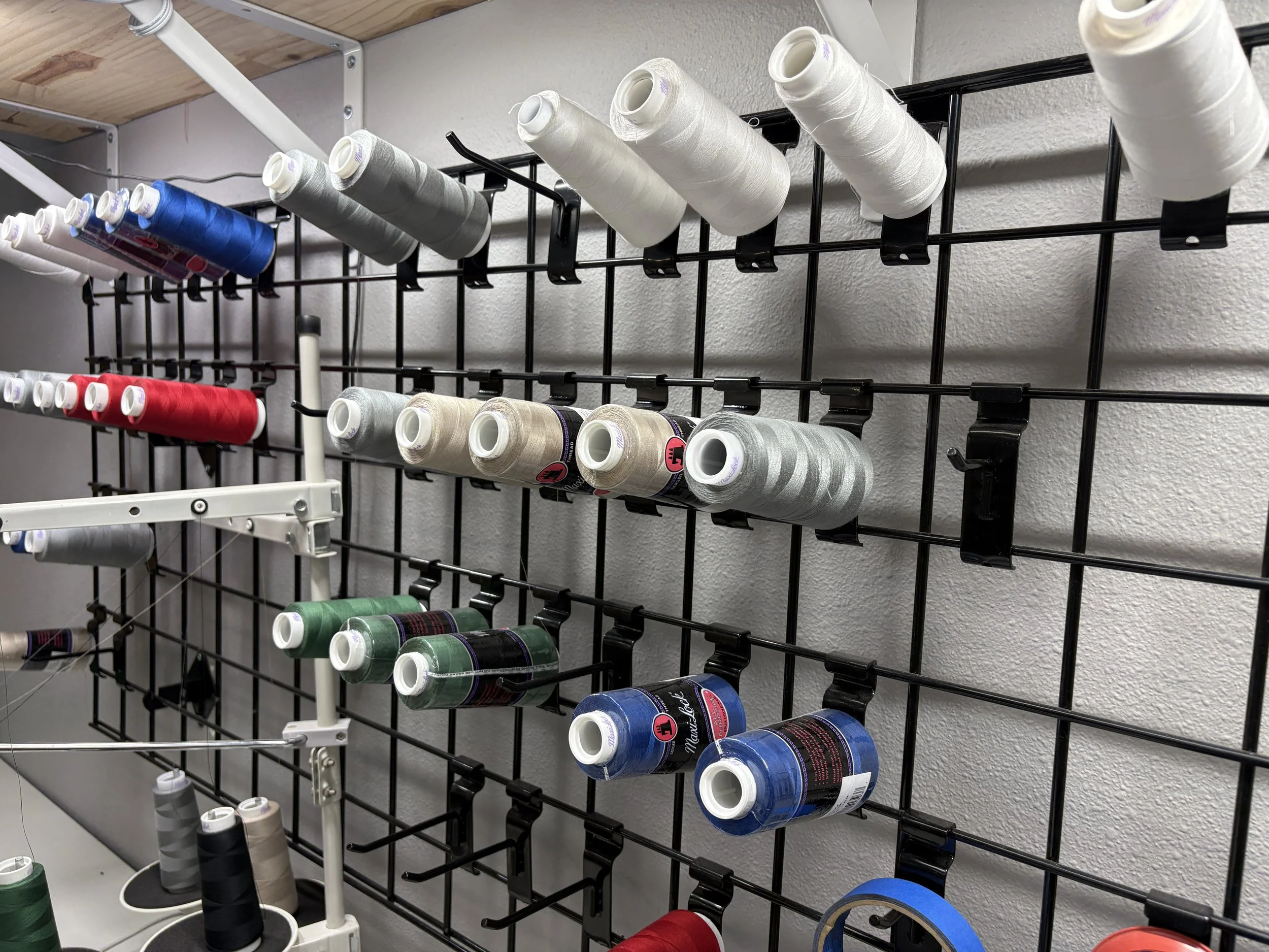 A black metal display rack holding various spools of thread in colors including blue, green, white, black, gray, and red. The rack is mounted on a wall with a wooden shelf and white wall in the background.