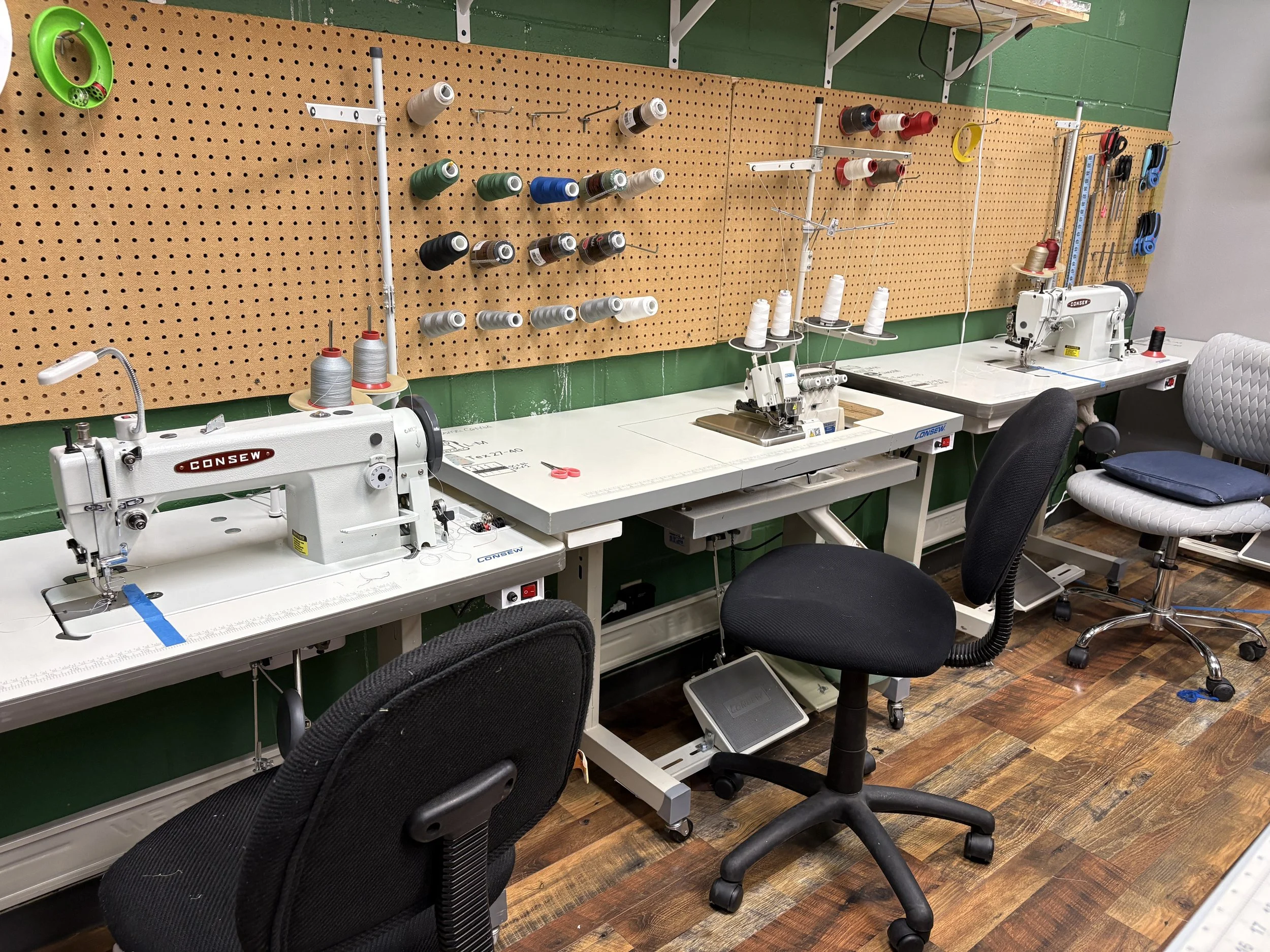 A workspace with multiple industrial sewing machines, sewing supplies, and chairs, with a large pegboard holding spools of thread and sewing tools.
