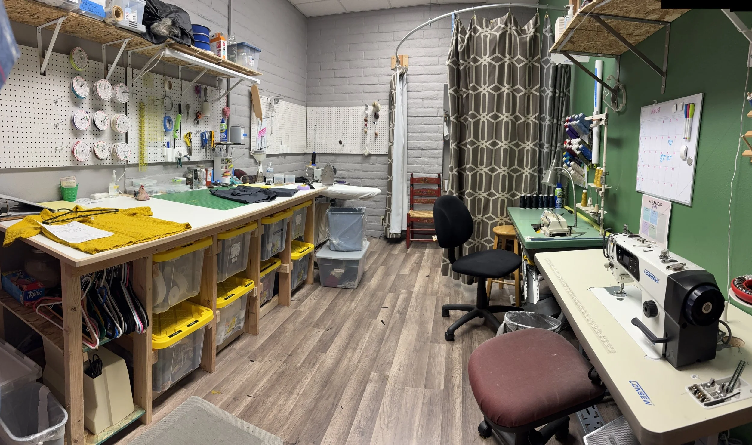 A well-organized craft room with workbenches, sewing machines, and supply storage. The room features a pegboard with tools, a curtain separating a workspace, and various crafting supplies on shelves and tables.