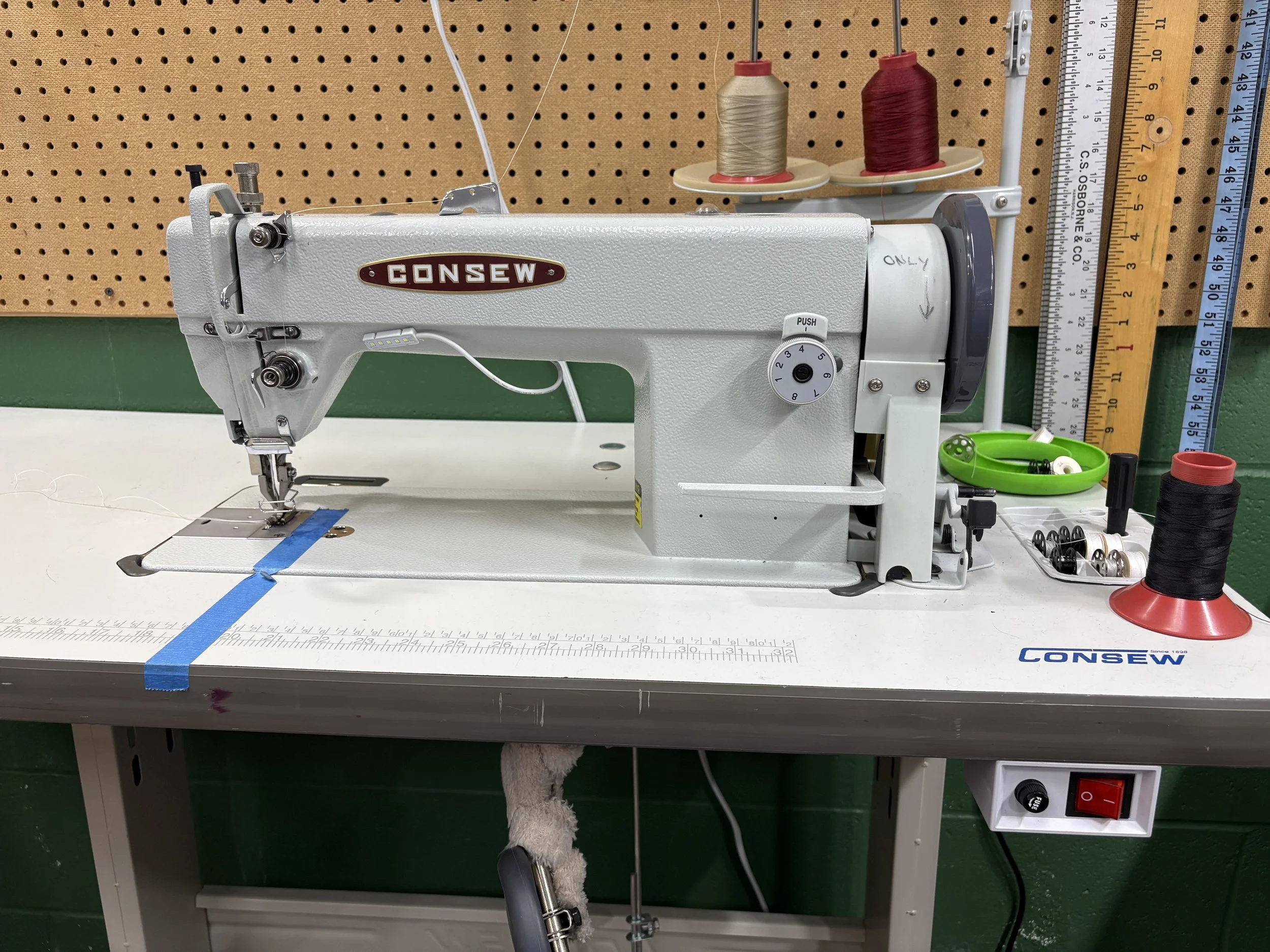 A sewing machine on a table in a workshop, with spools of thread, rulers, and other sewing tools nearby, and a pegboard in the background.