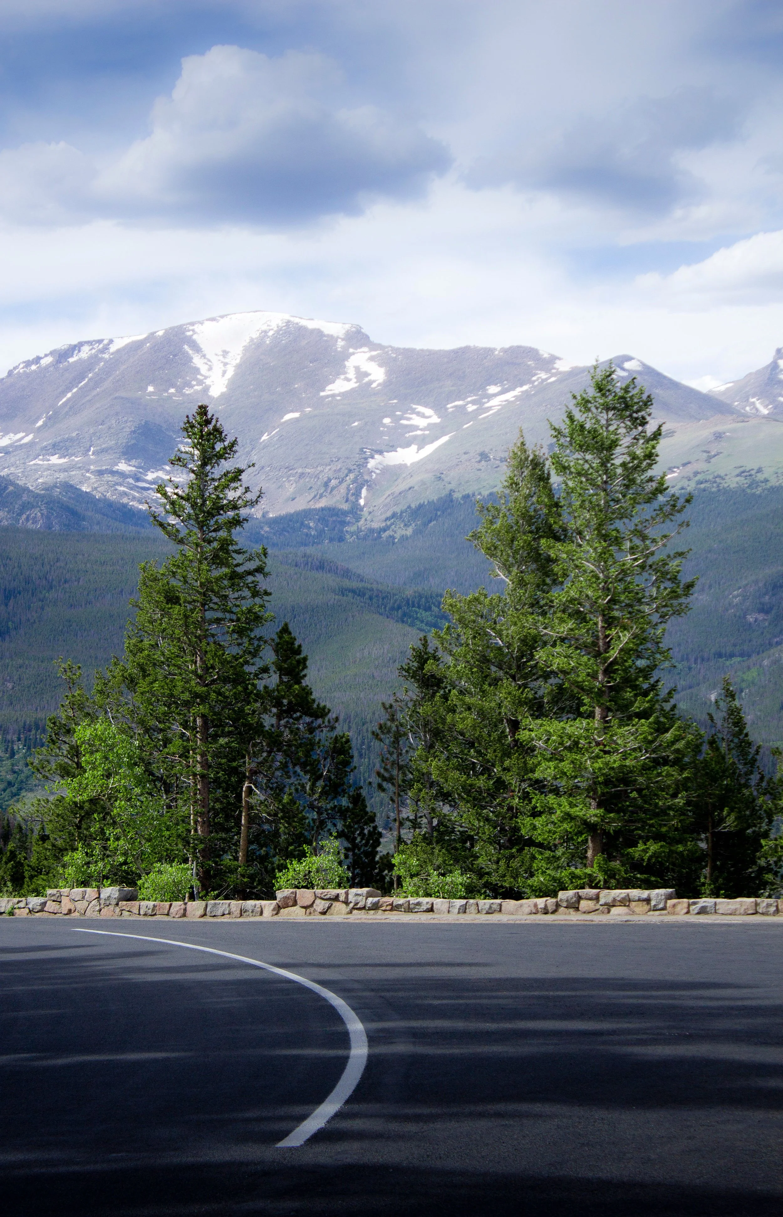 A winding mountain road with a stone barrier on the side, green trees, and snow-capped mountains in the background under a partly cloudy sky.