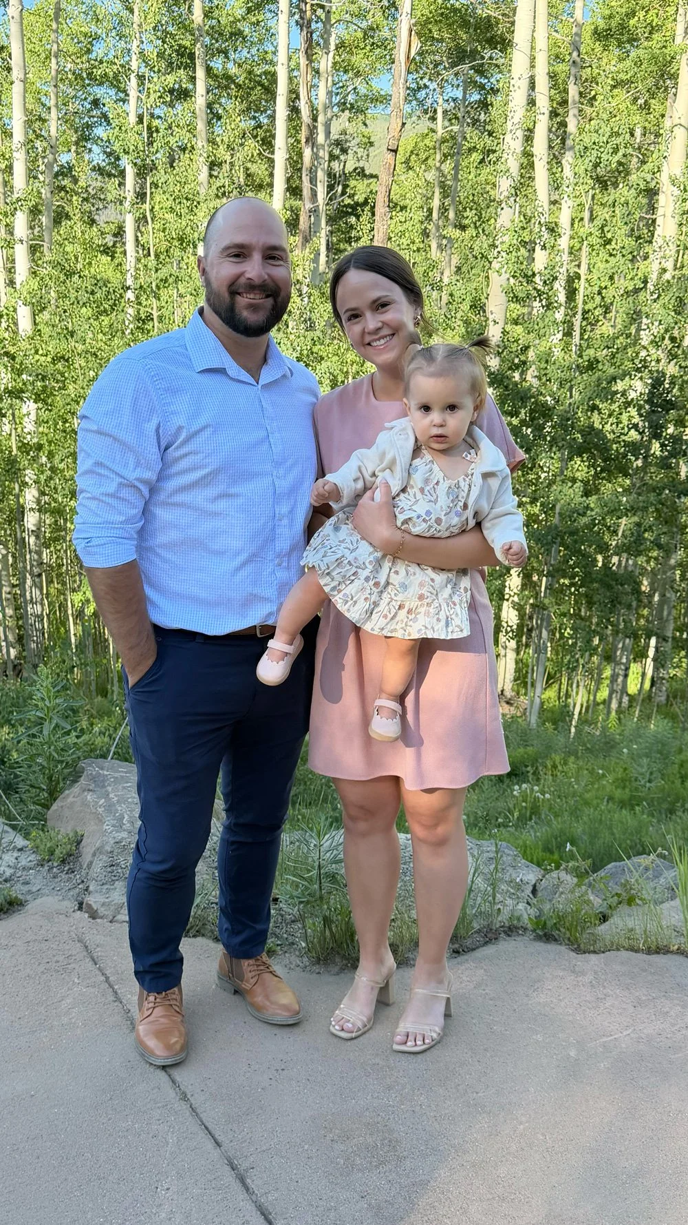 A family of three standing outdoors in a forested area. The man is on the left, wearing a light blue checkered shirt and dark pants. The woman is in the middle, holding a young girl, wearing a pink dress and beige heels. The girl has twin pigtails and is wearing a floral dress with a light jacket, white shoes, and socks. They are smiling and posing for the photo.