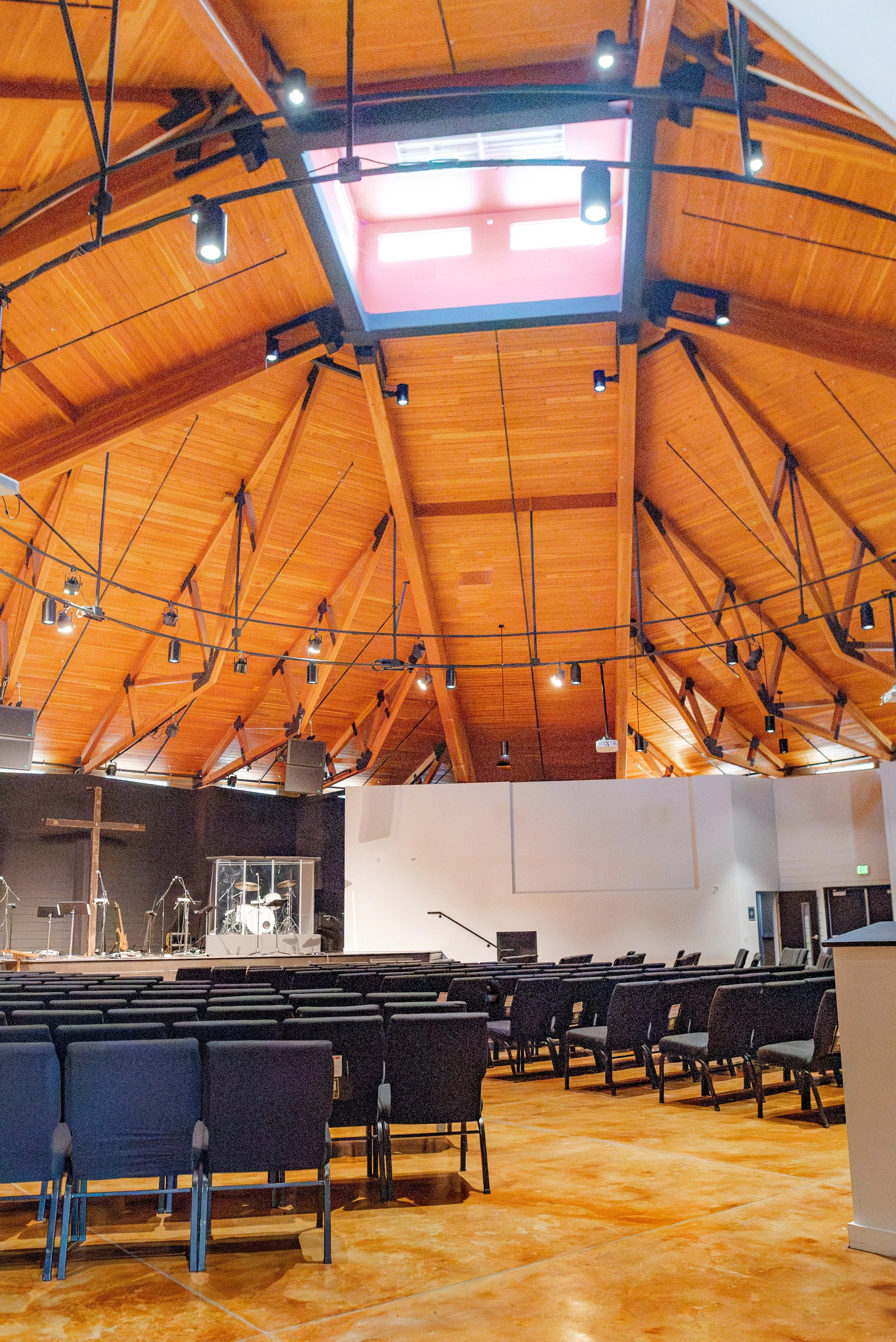 Interior of a church or auditorium with wooden ceiling, rows of black chairs, a stage with musical instruments, a cross, and a large screen hanging from the ceiling.