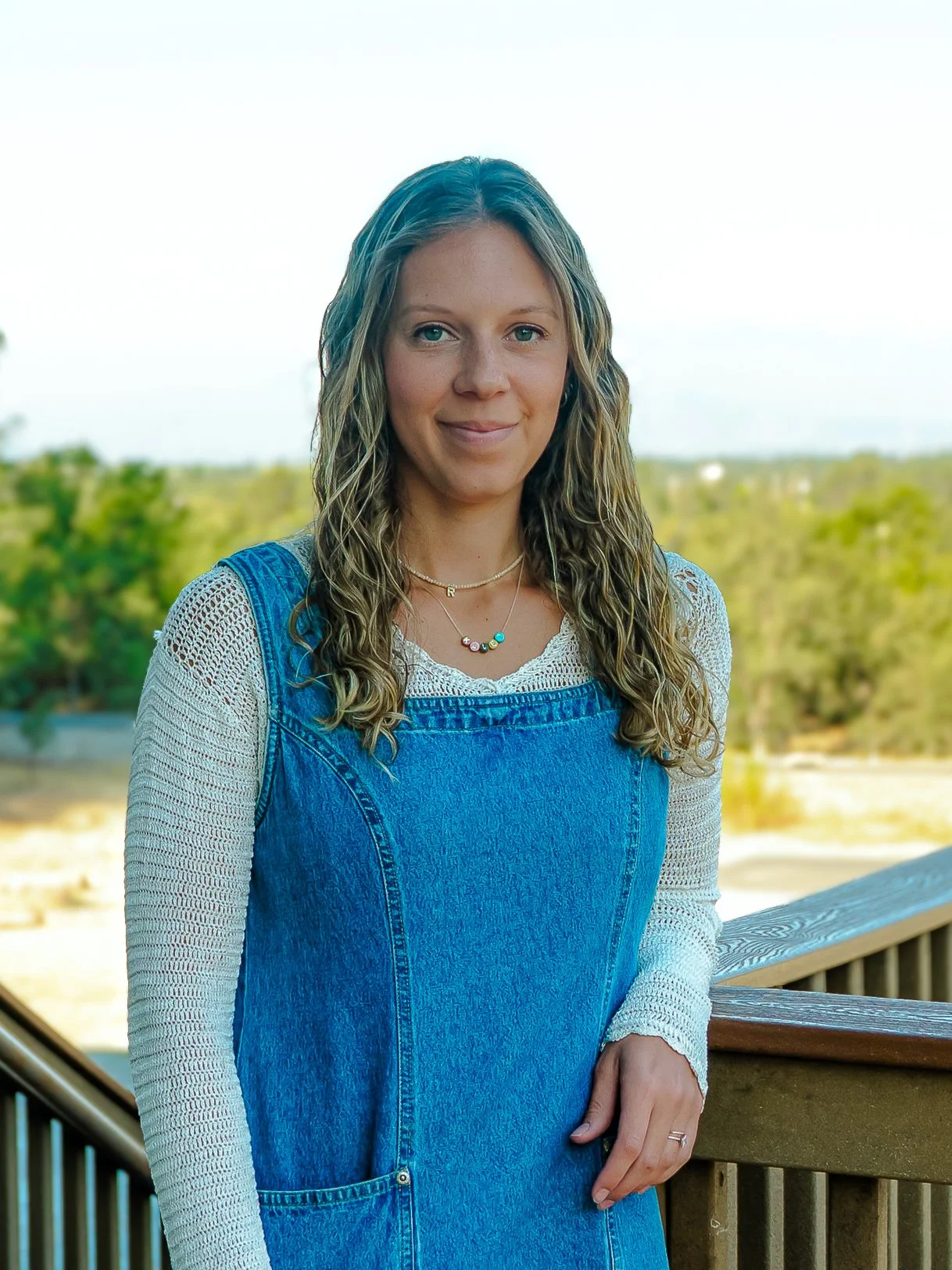 A young woman with long wavy blonde hair and light skin standing outdoors on a wooden deck, wearing a white long-sleeve top underneath blue denim overalls, with necklaces and a ring on her finger. The background features trees and a clear sky.
