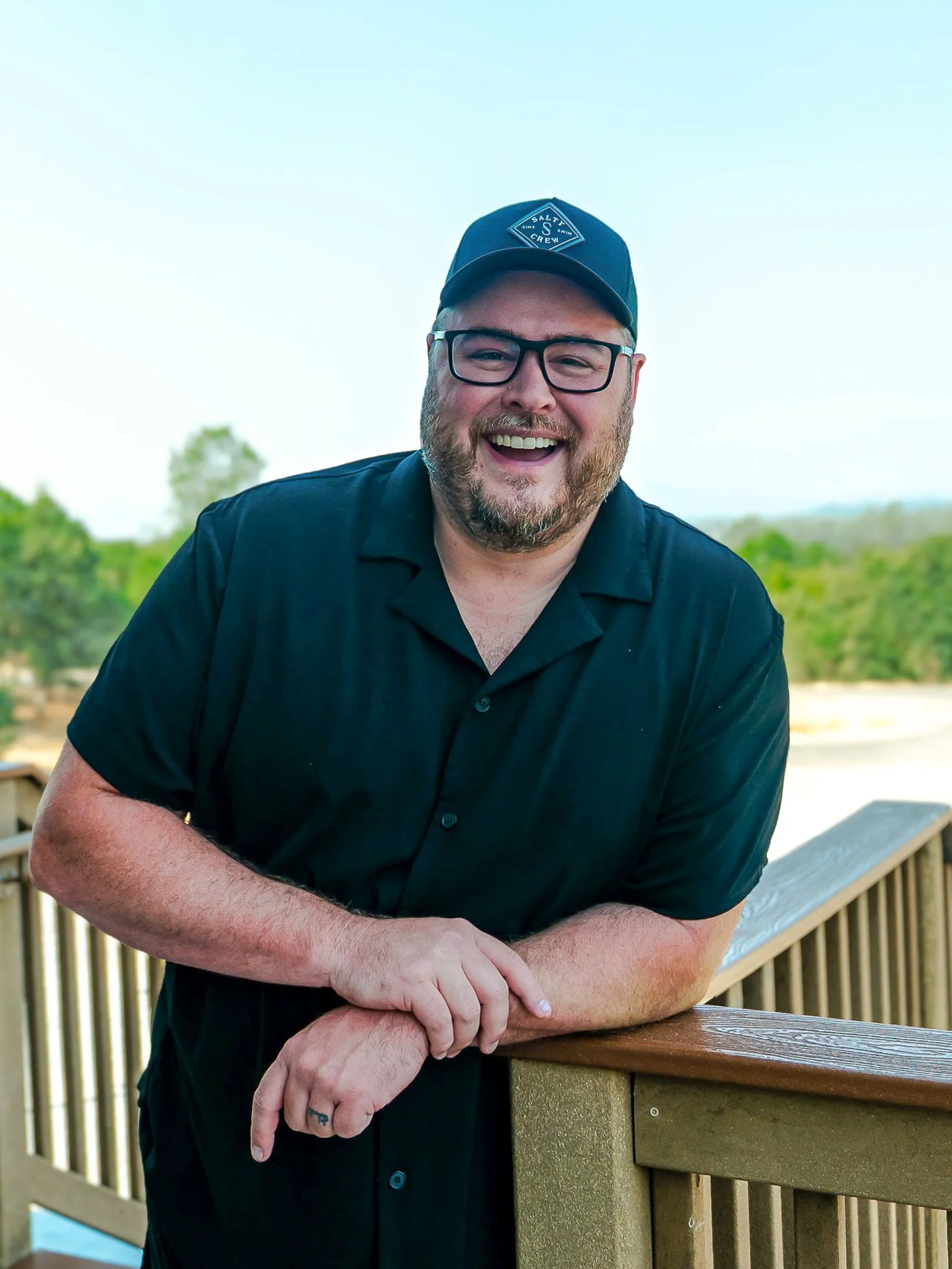 A smiling man with glasses and a beard leaning on a wooden railing outdoors, wearing a black shirt and a navy baseball cap with a logo.