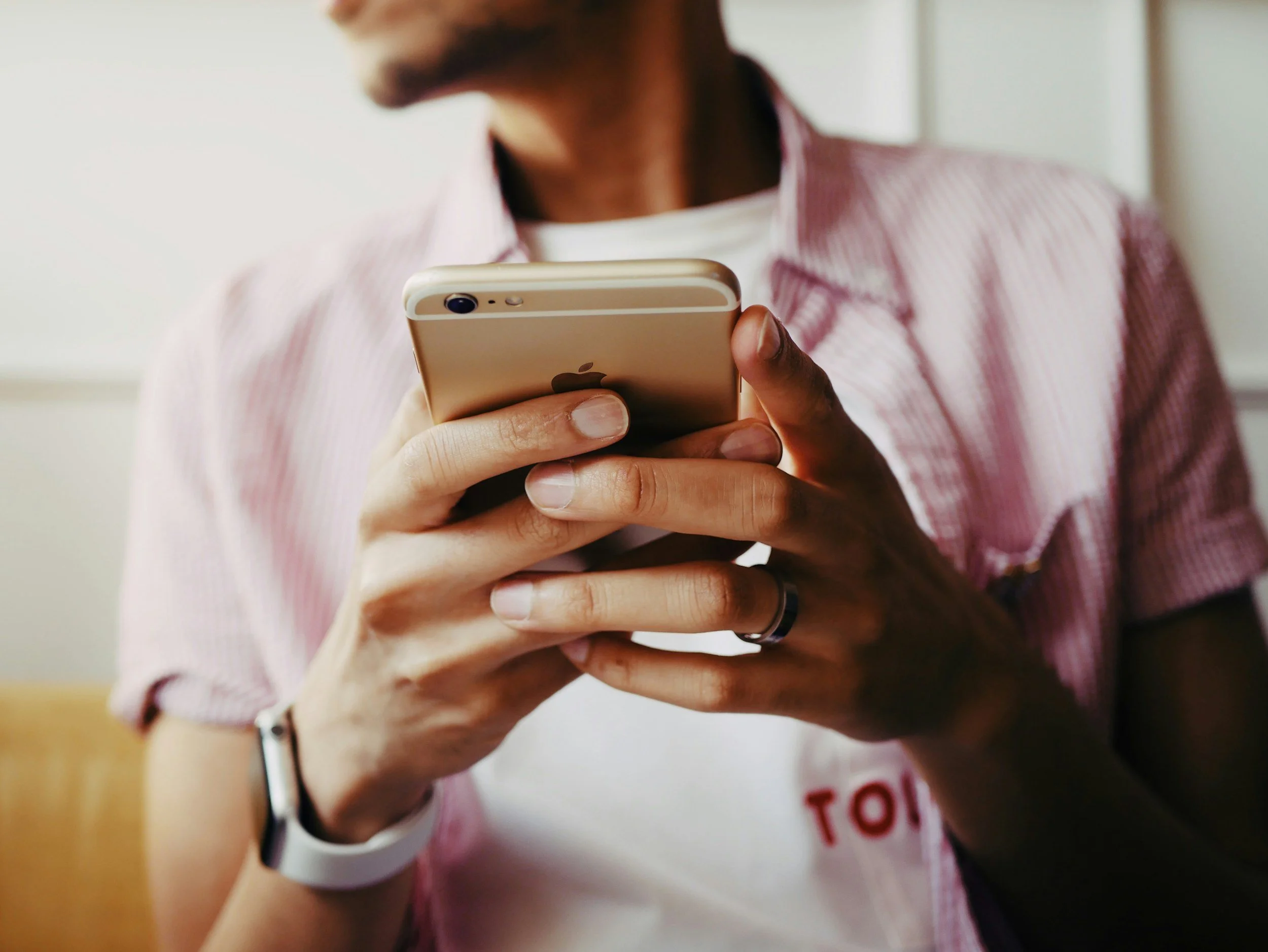 Person holding a smartphone with a beige back and black camera, wearing a wristband and bracelet, and dressed in a pink striped shirt with a white T-shirt underneath.