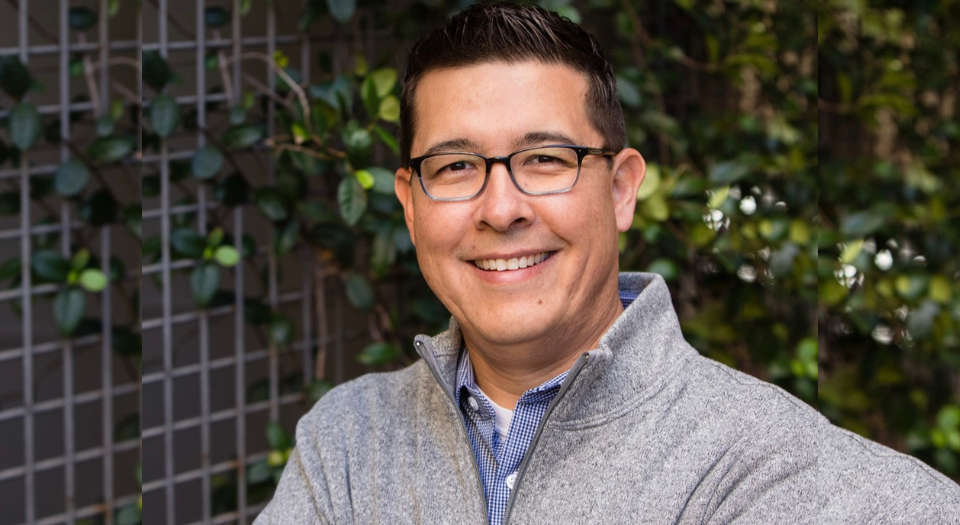 A smiling man with glasses wearing a gray zip-up sweater and a blue collared shirt, standing outdoors in front of a leafy green background.