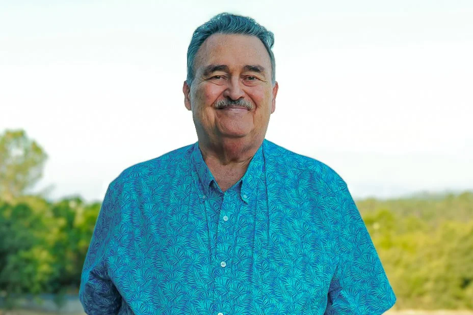 Smiling middle-aged man with gray hair and mustache wearing a blue patterned button-up shirt outdoors with a blurred green landscape in the background.