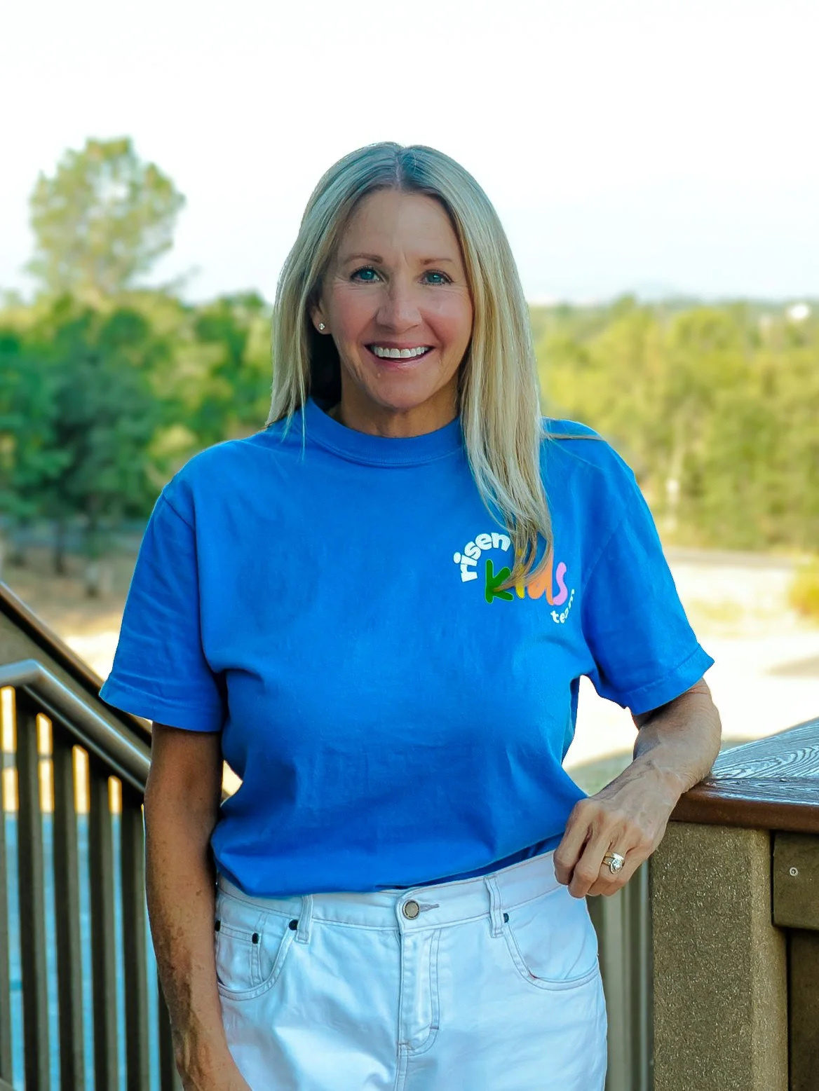 A smiling woman with blonde hair wearing a blue T-shirt and white shorts standing outdoors on a deck with a railing, with trees and a clear sky in the background.